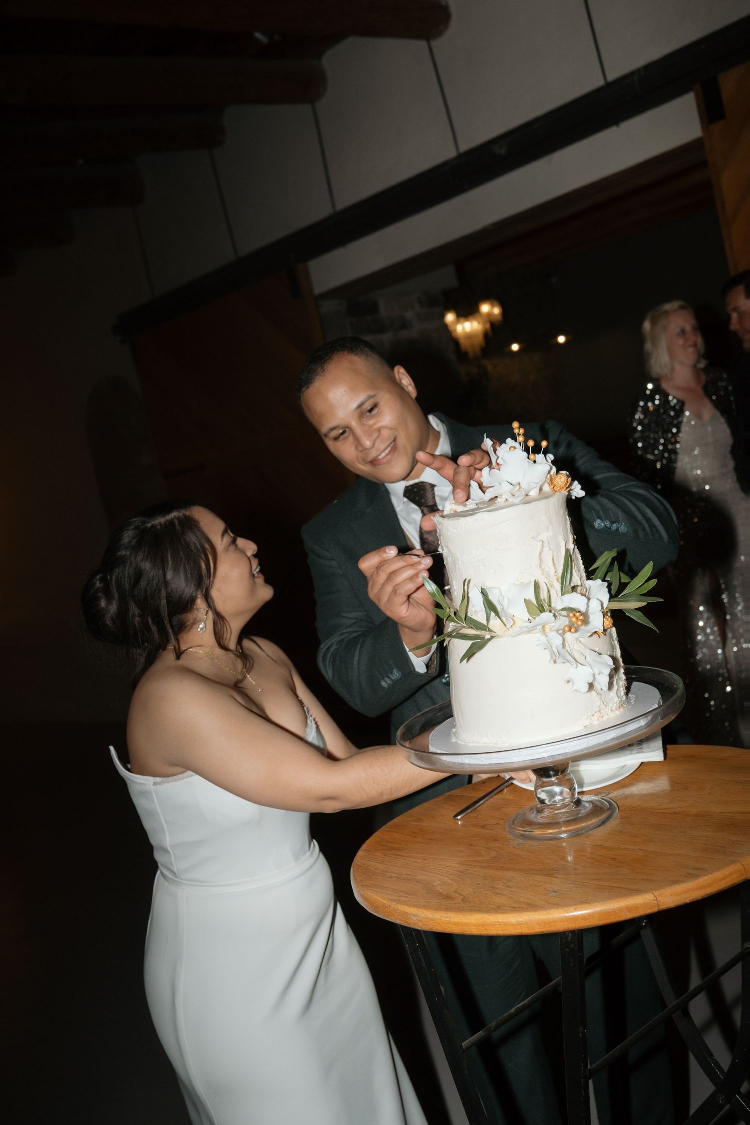 Bride and groom cutting their elegant two-tier white wedding cake decorated with greenery and flowers.