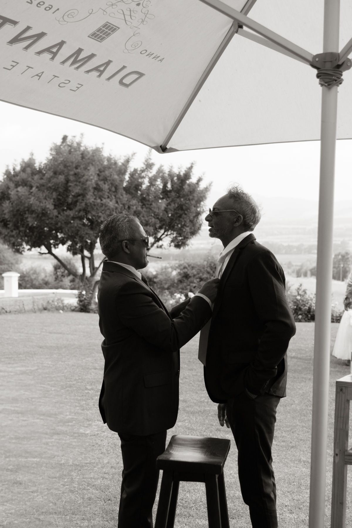 Two men in suits share a handshake under a Diamond Estate umbrella in a black and white wedding photo.