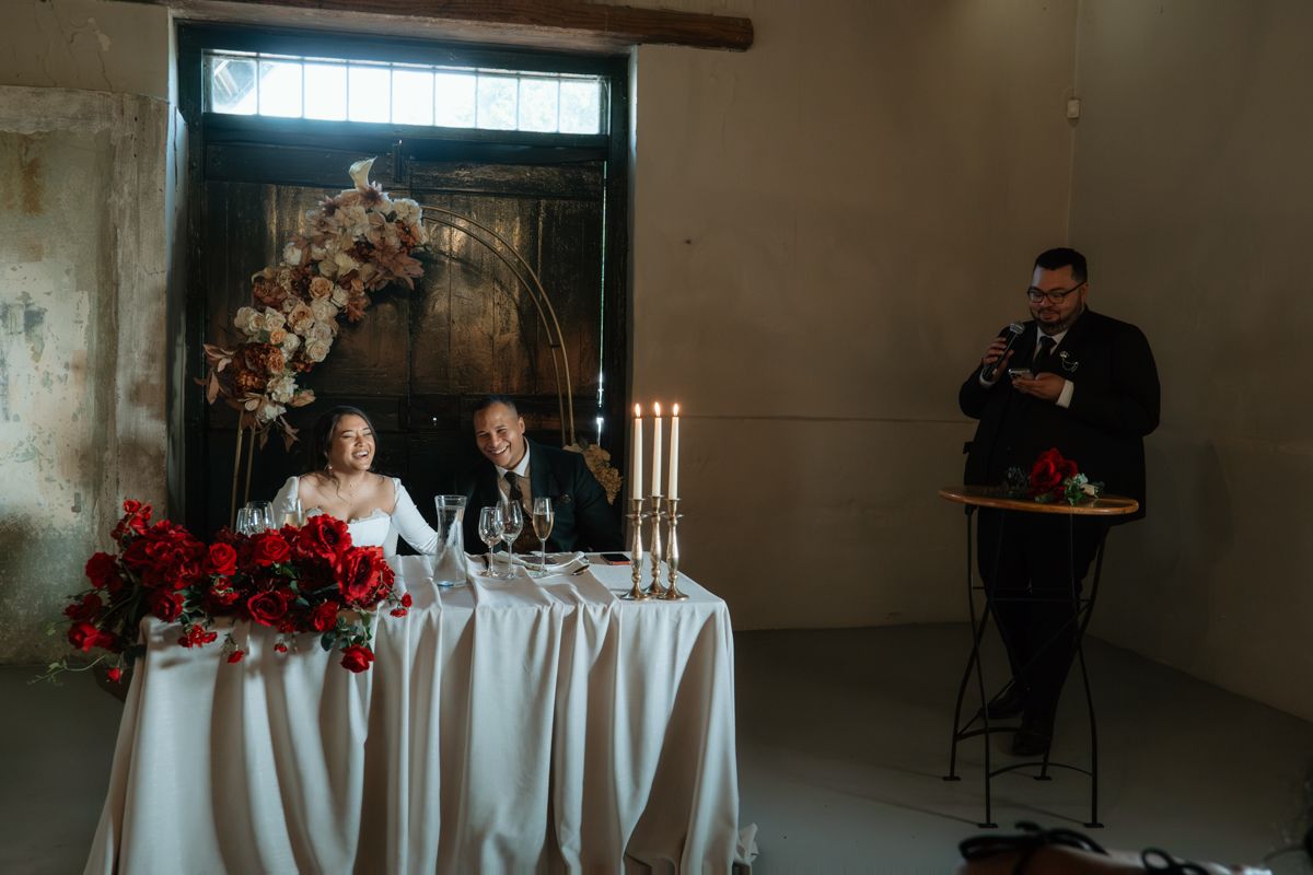 Bride and groom sit at sweetheart table with red floral arrangements and candles at wedding reception.