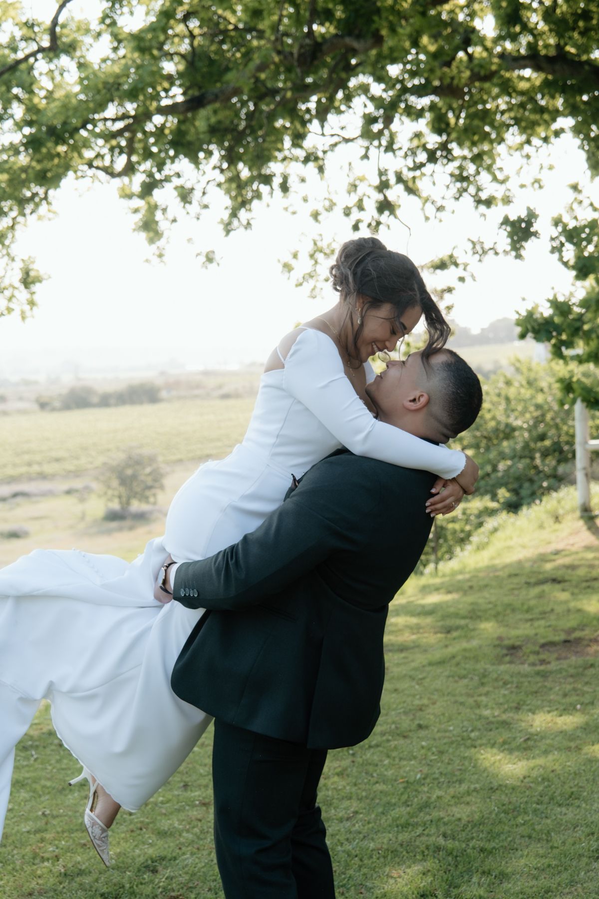 Groom lifting smiling bride in white gown under tree at golden hour outdoor wedding portrait.