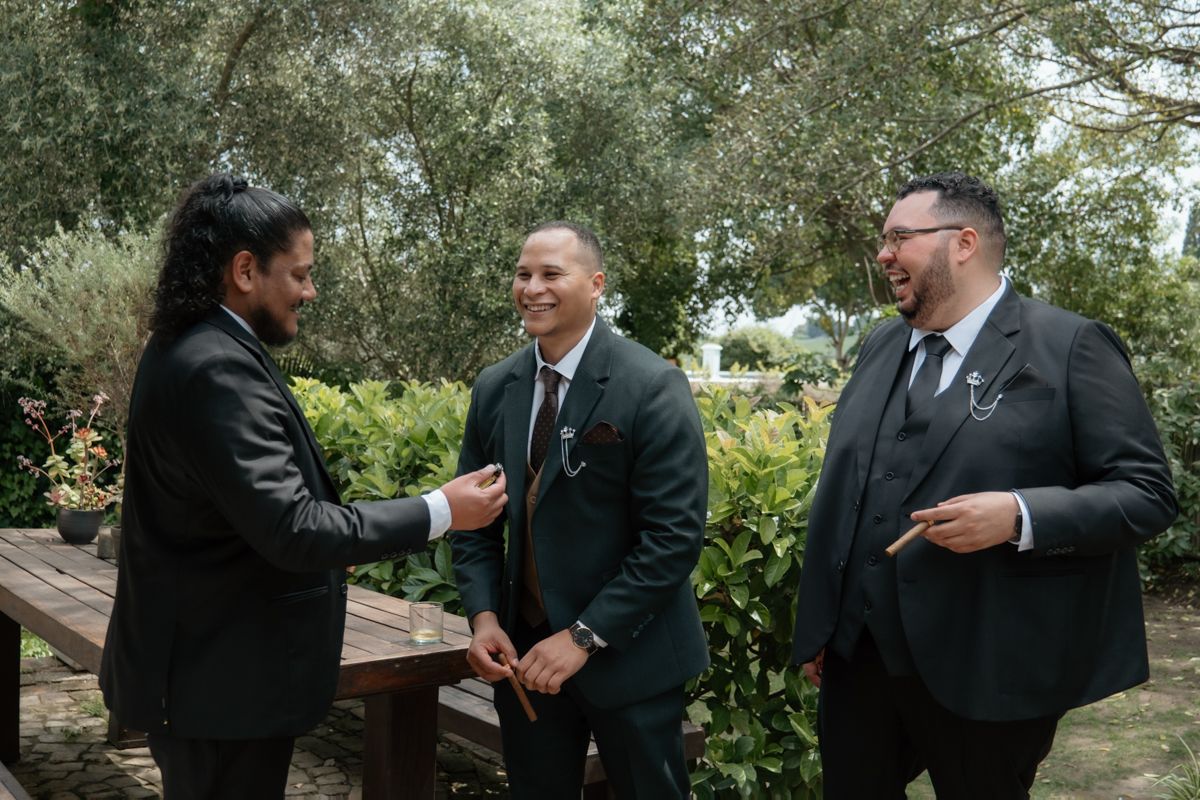 Three groomsmen in black suits laughing together outdoors in a lush garden before the wedding.