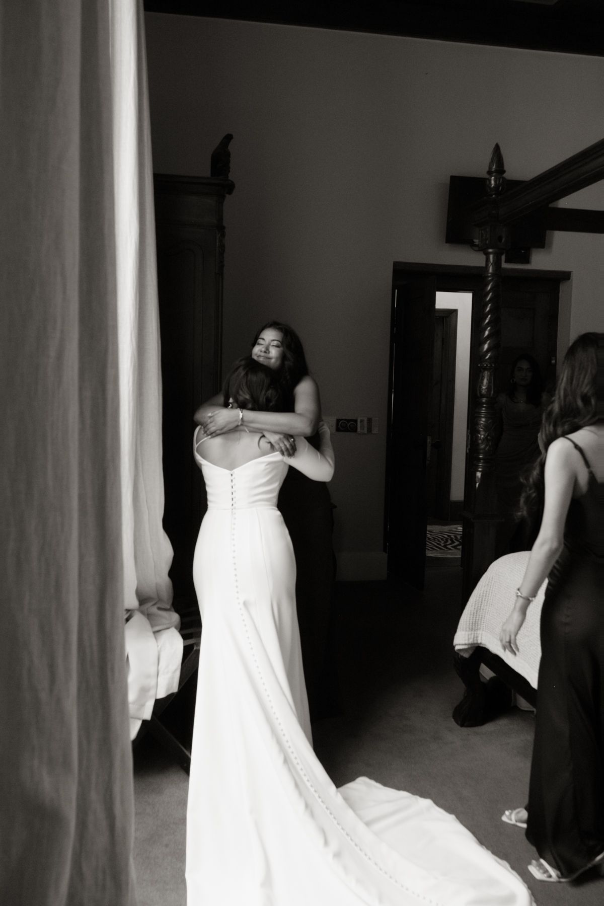 Black and white photo of bride in fitted white gown with train being embraced in a room before wedding ceremony.
