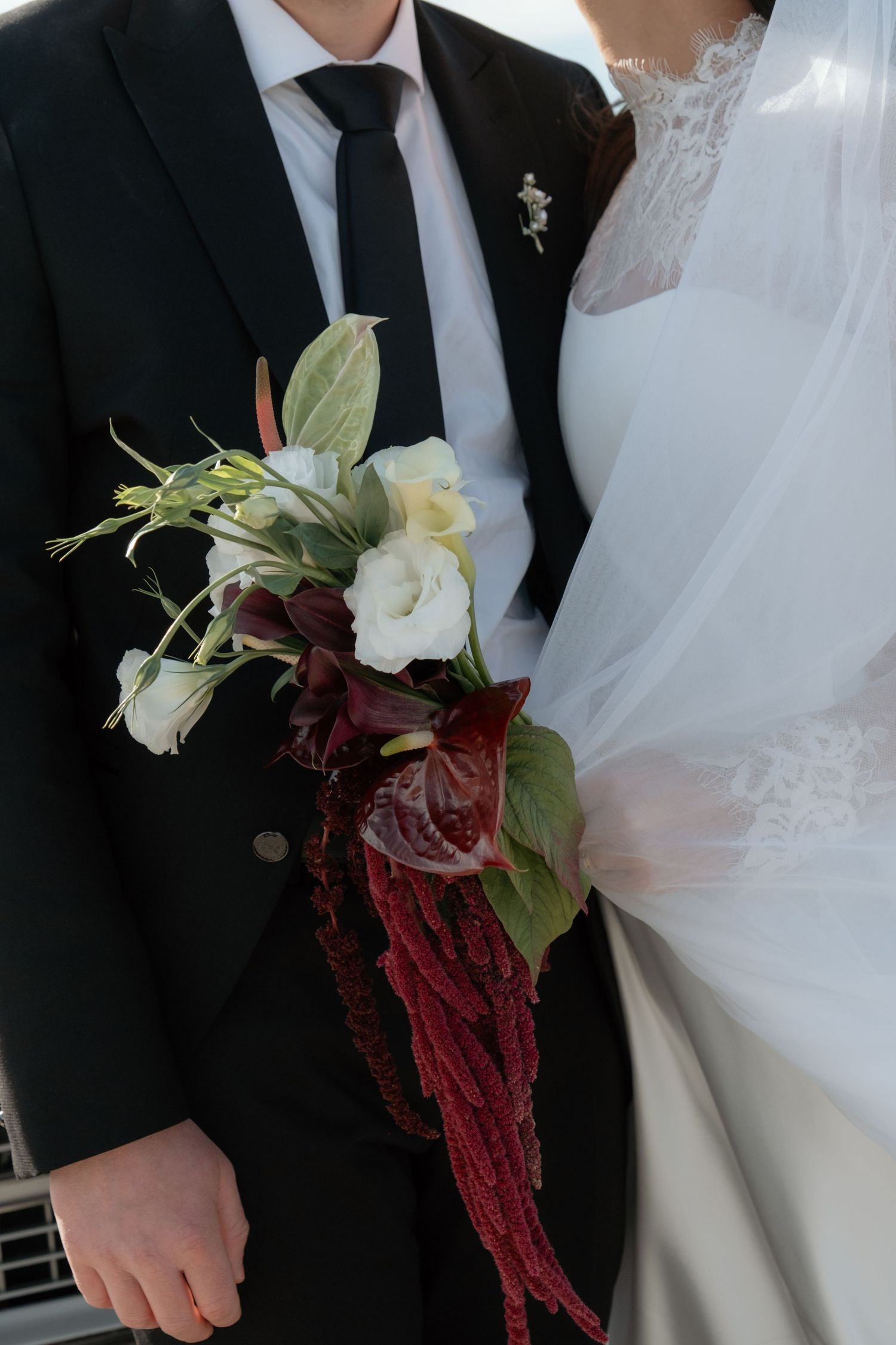 Bride holding a cascading wedding bouquet with white roses and deep red amaranthus beside groom in black suit.