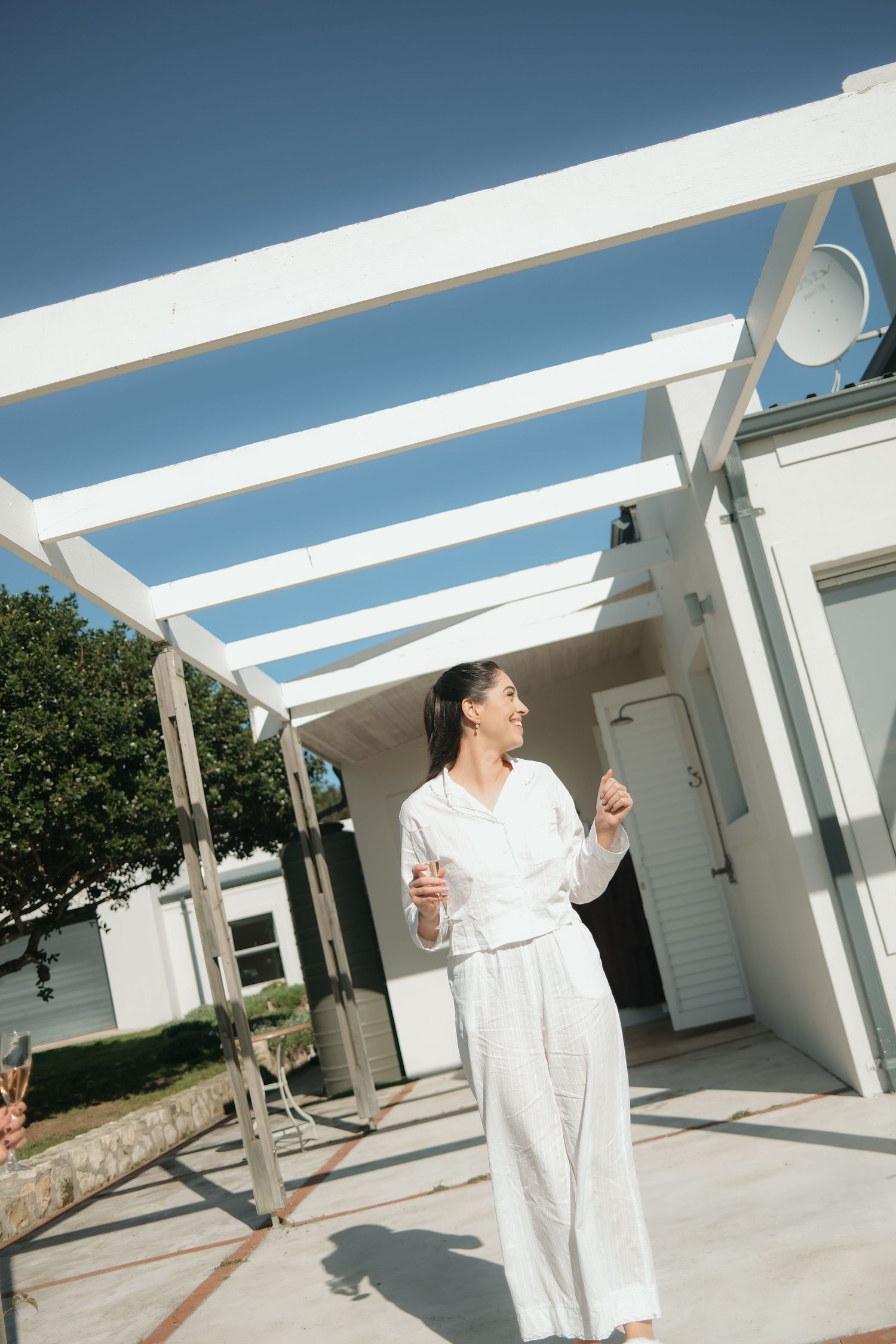 Bride in white robe holding champagne glass outdoors under pergola on sunny wedding day.