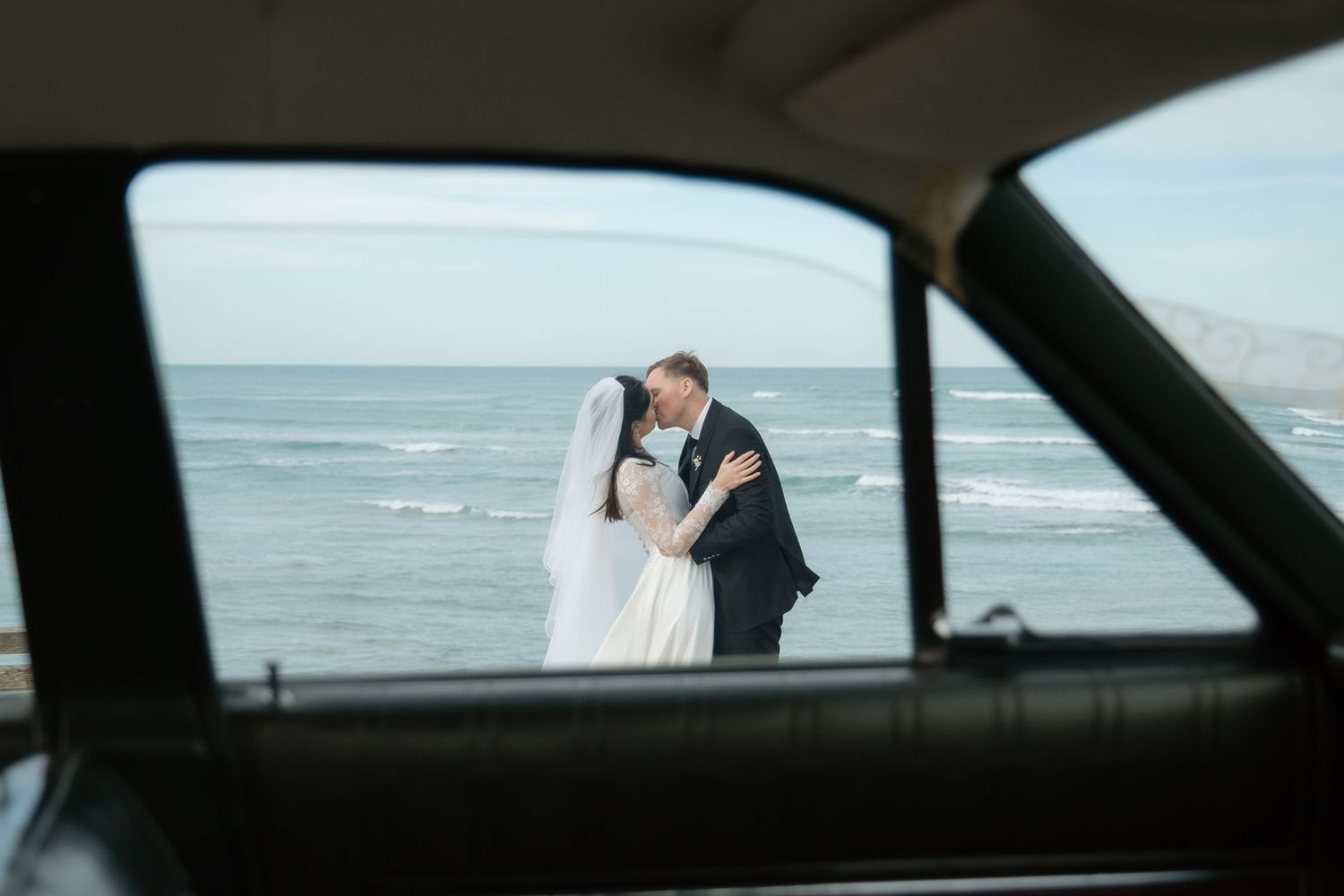 Bride and groom kissing by the ocean, framed through a vintage car window on their wedding day.