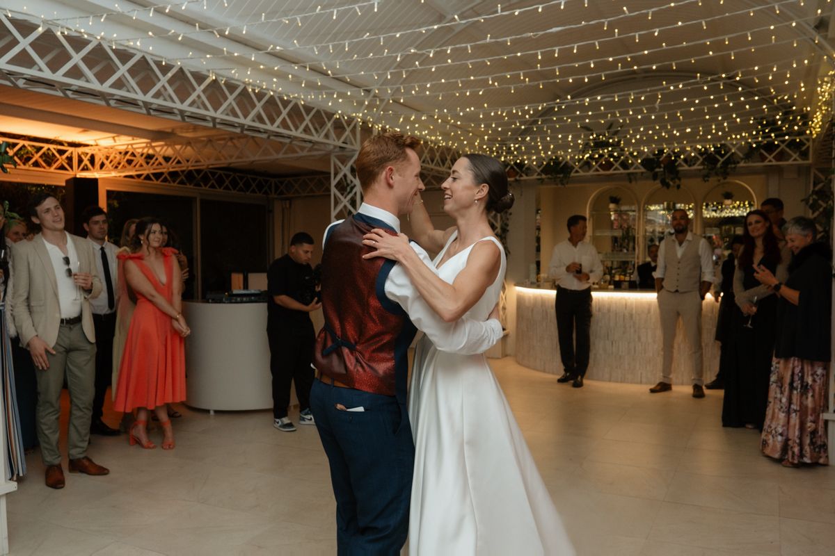 Bride and groom sharing their first dance under fairy lights while wedding guests look on.