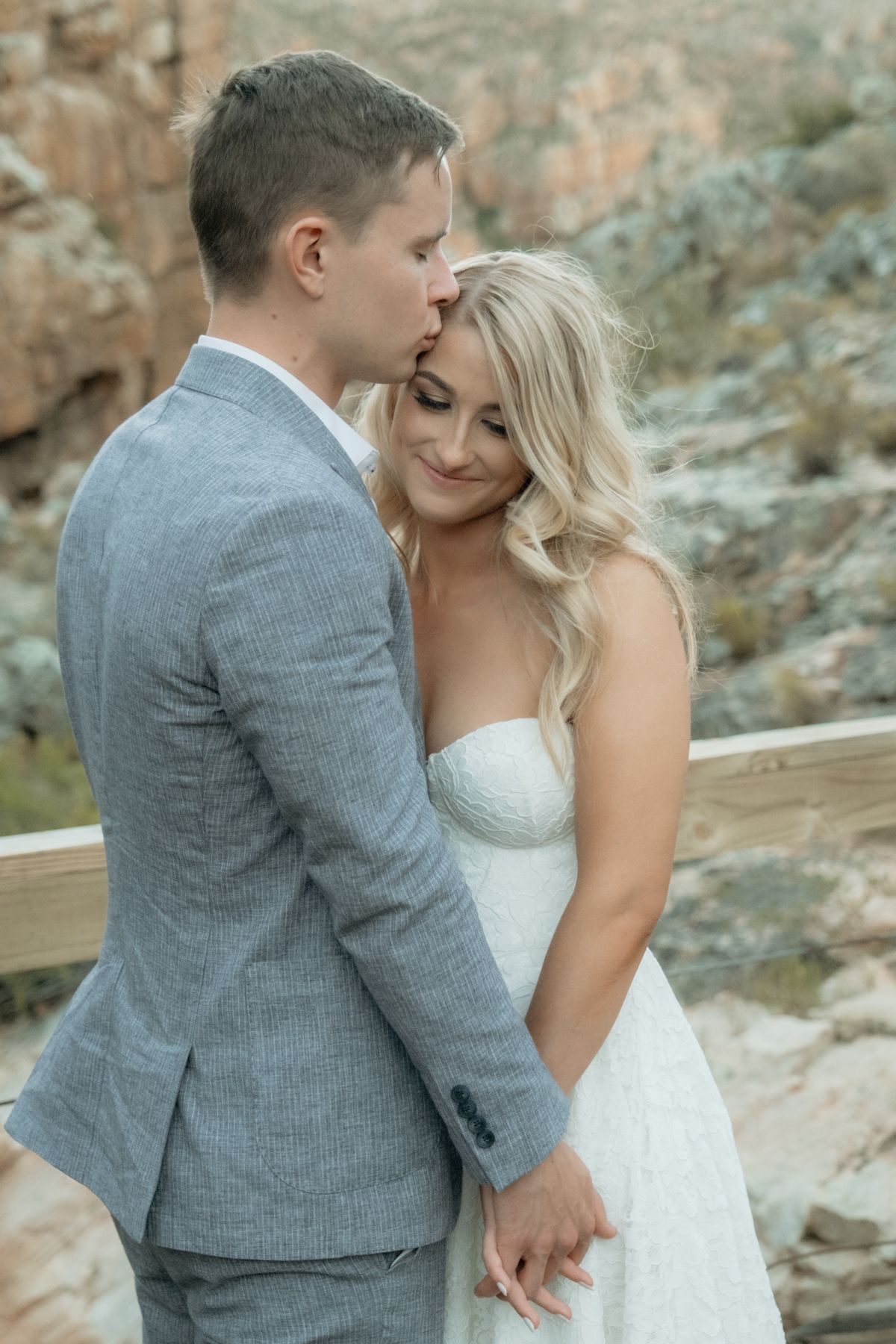 Groom in grey suit tenderly kissing bride on forehead while holding hands on a rustic wooden bridge.
