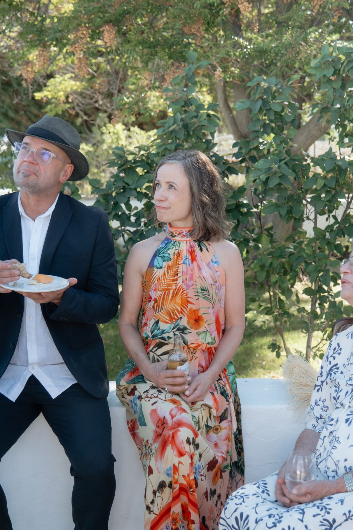 Wedding guests in colorful floral dress and black suit enjoy outdoor reception under green trees.