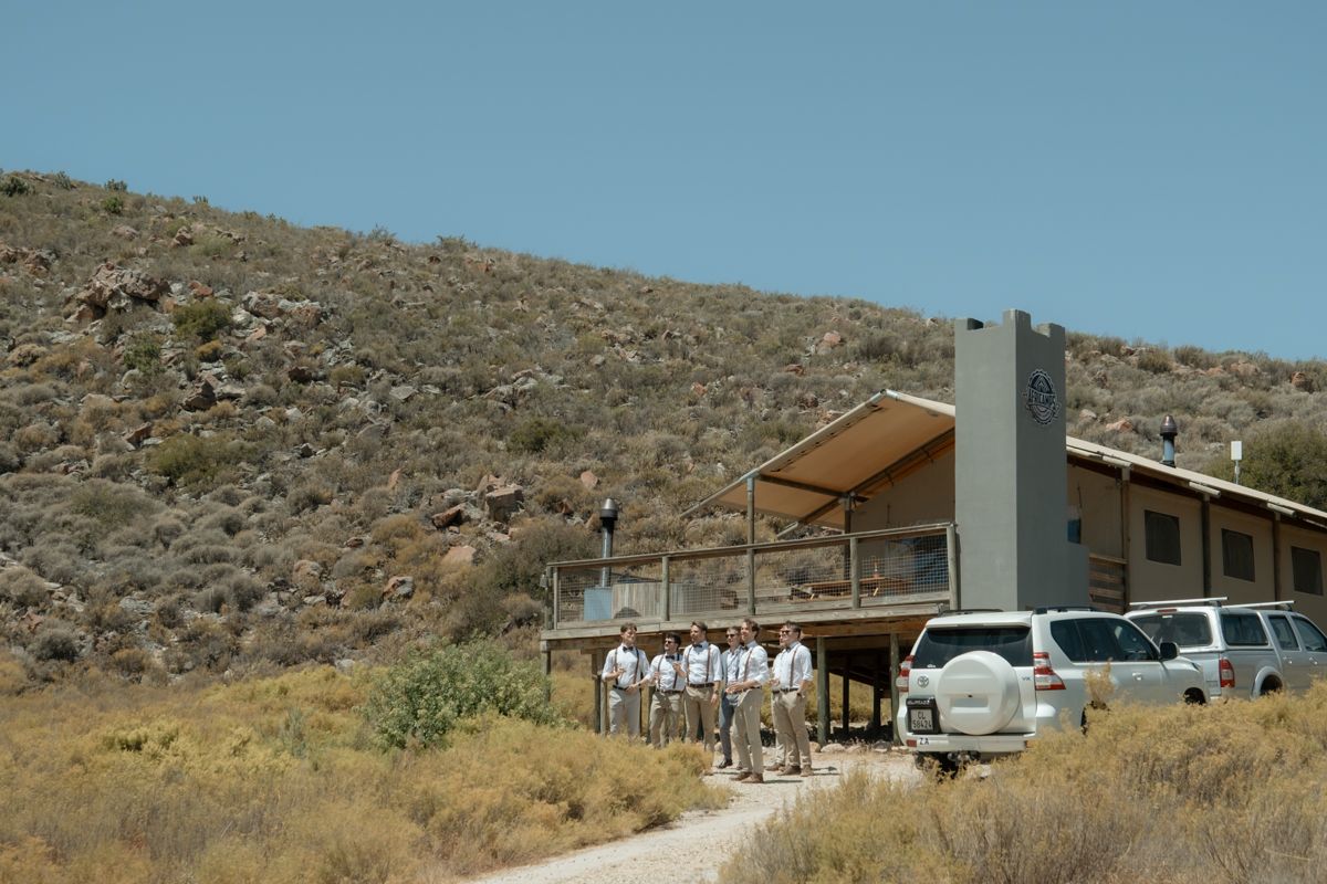 Wedding party group standing on deck of modern building nestled against dramatic desert hillside with parked vehicles.