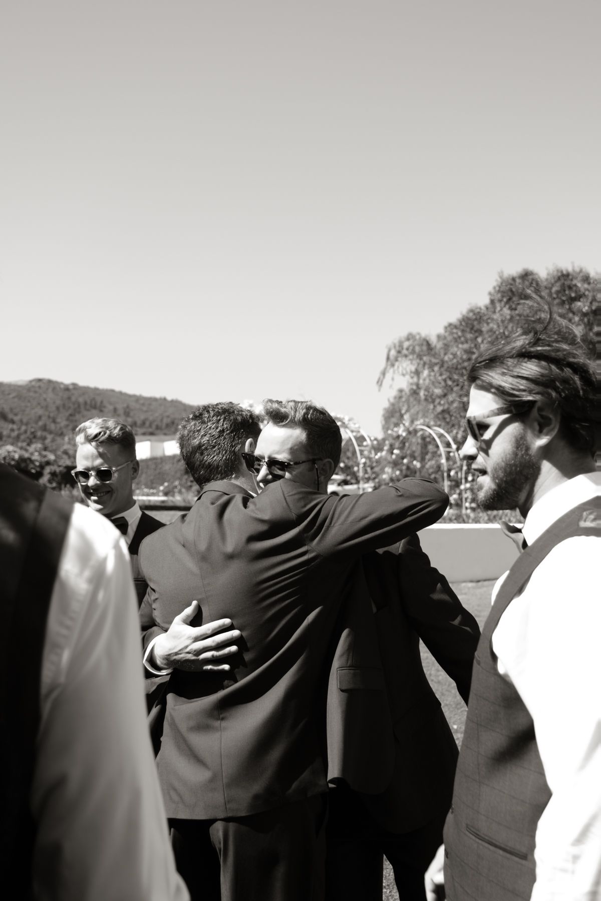 Black and white photo of groomsmen embracing and congratulating each other at an outdoor wedding.