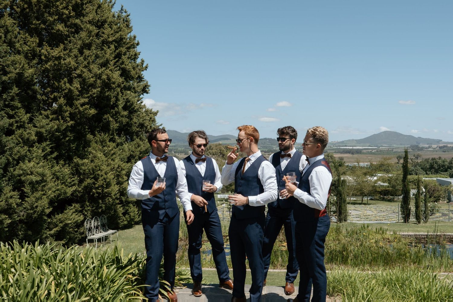 Five groomsmen in navy vests and bow ties celebrate outdoors with drinks against a scenic countryside backdrop.
