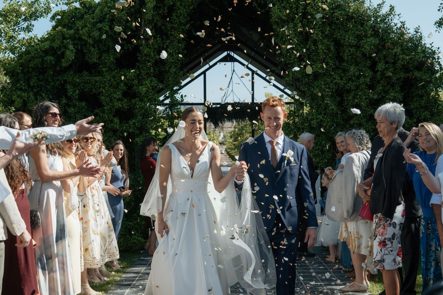 Newlyweds walk through confetti shower as smiling guests celebrate their outdoor wedding ceremony exit.