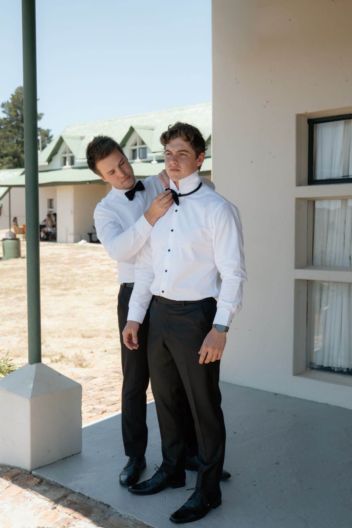 Groomsman helping groom adjust black bow tie outdoors before wedding ceremony in formal tuxedo attire.