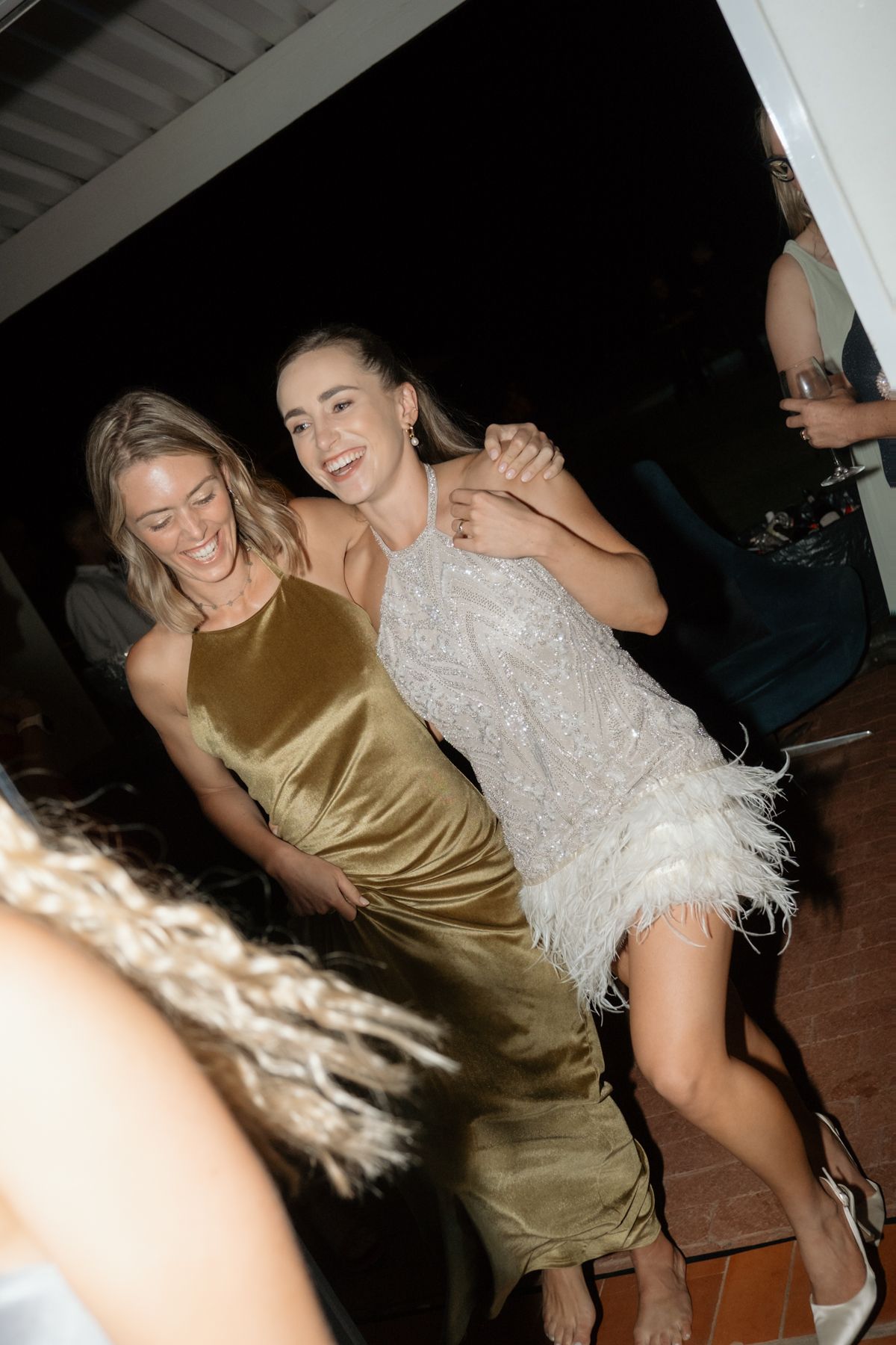 Bride in sequin feather dress dancing joyfully with guest in gold velvet gown at reception.
