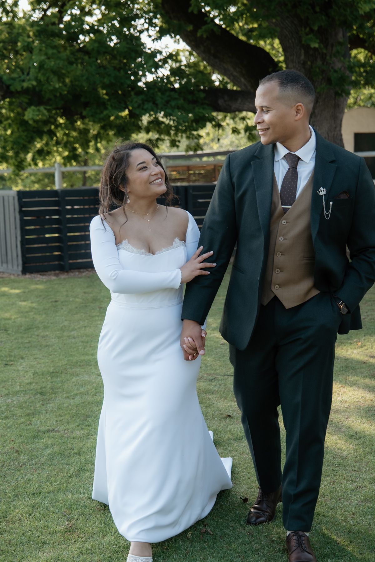 Bride in white gown and groom in dark green suit walk hand in hand smiling at each other outdoors.