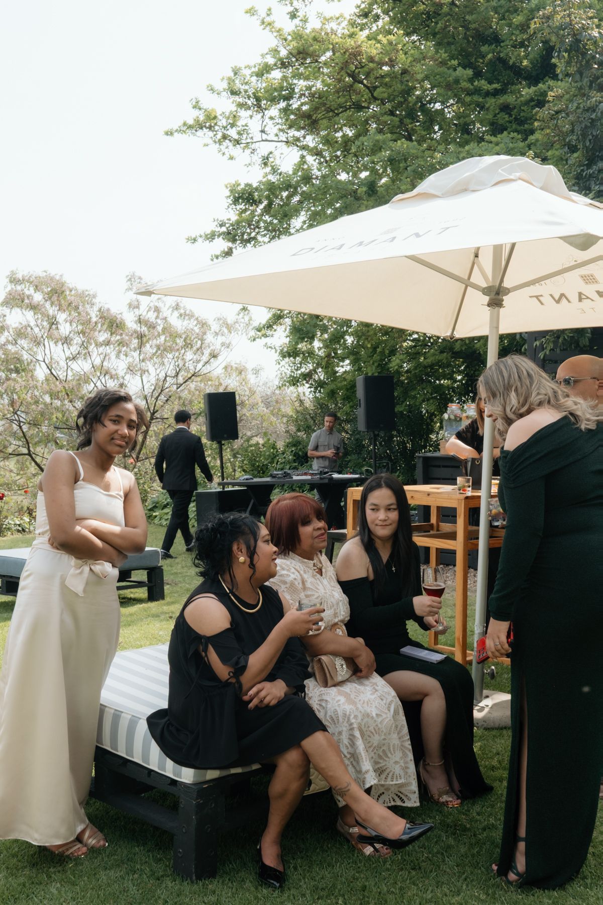 Bride in white gown mingles with elegantly dressed wedding guests seated outdoors under white umbrella.