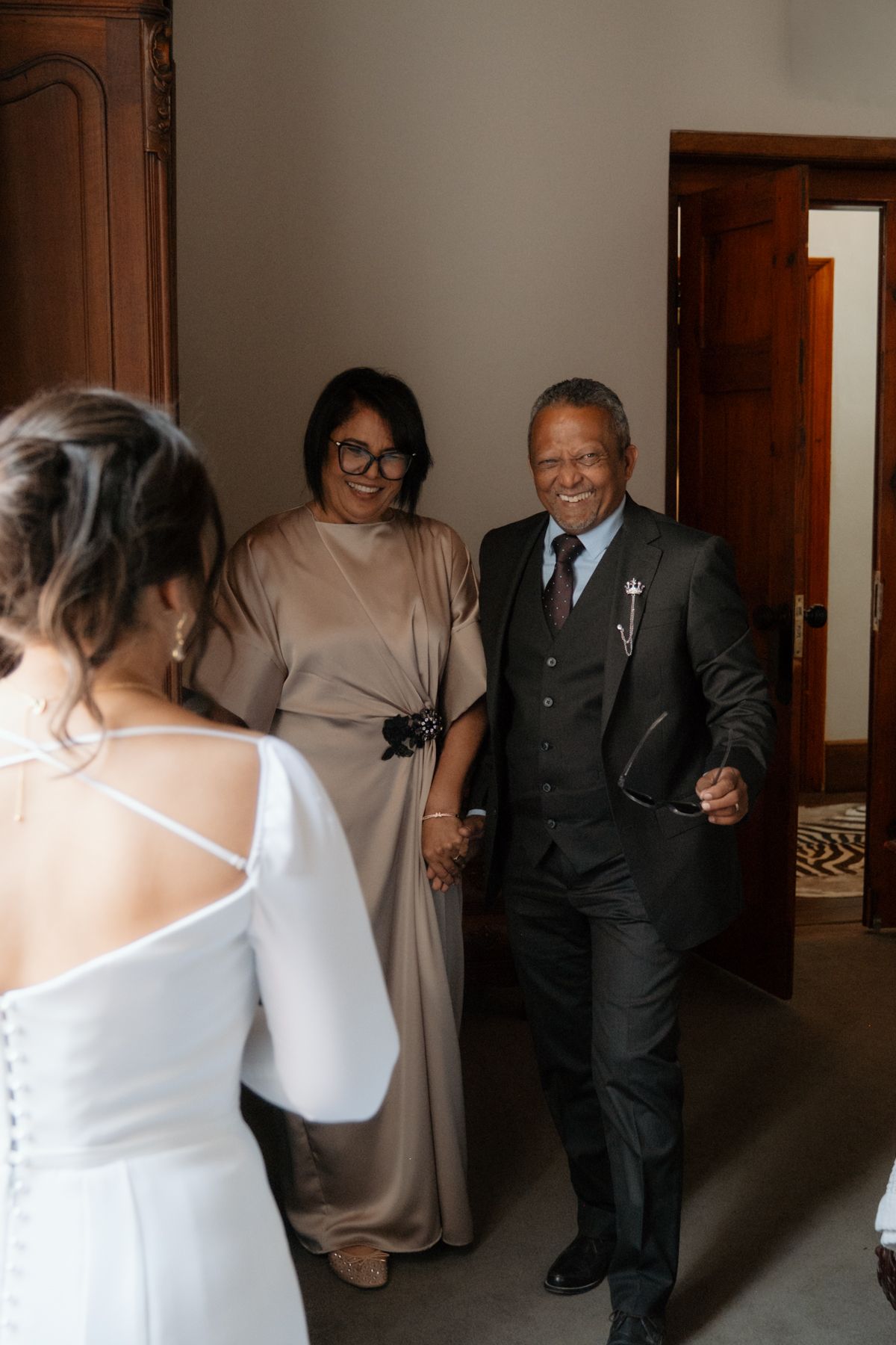 Proud parents smile as they enter room to see bride in white backless wedding gown during wedding preparations.
