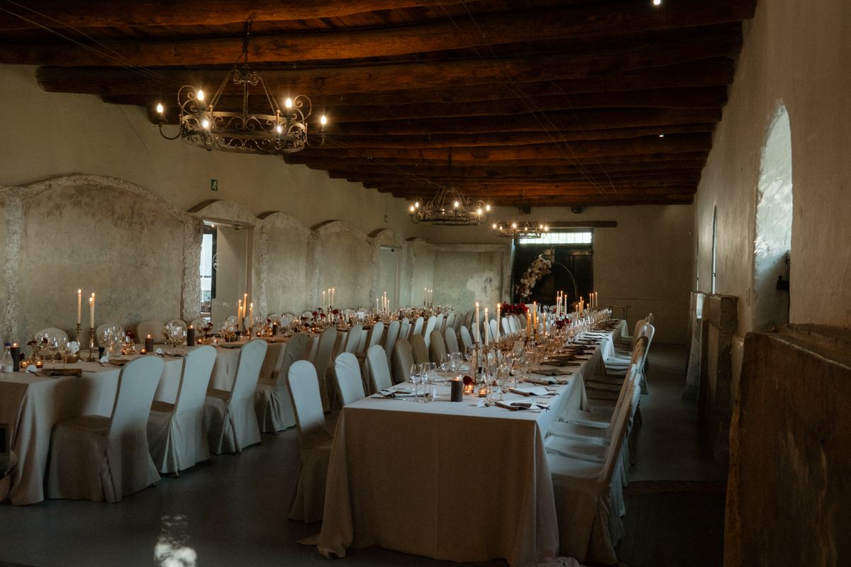 Elegant wedding reception hall with long tables, white chairs, candlelight, and rustic wooden ceiling.
