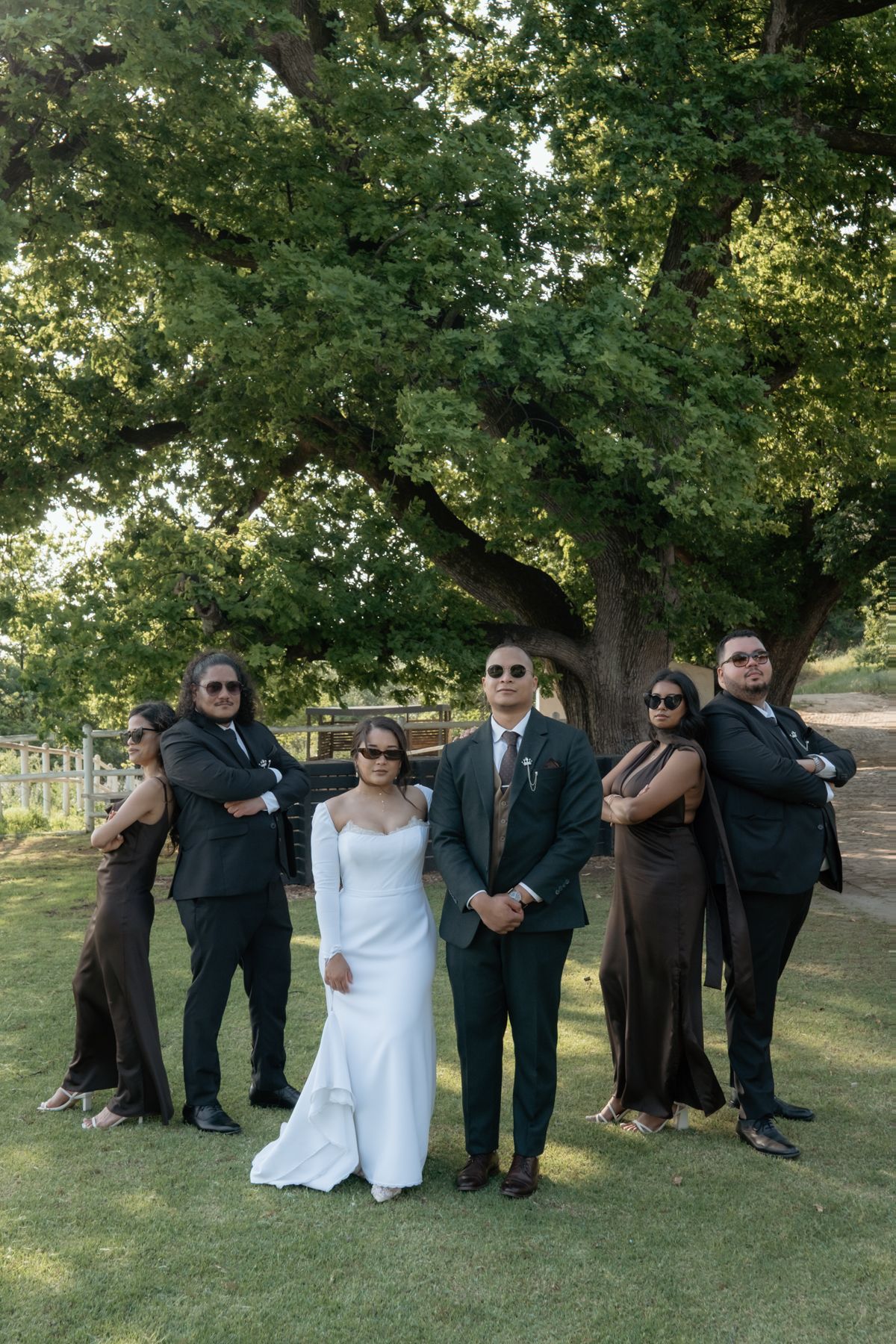 Wedding party posing coolly with sunglasses under a large green tree outdoors.