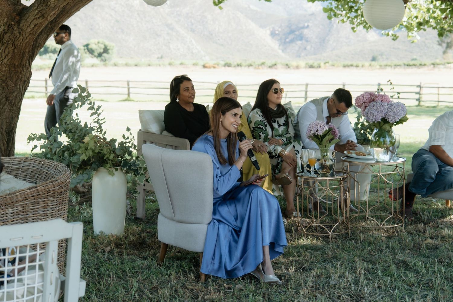 Woman in blue dress seated outdoors at an elegant garden party with guests and floral decorations nearby.