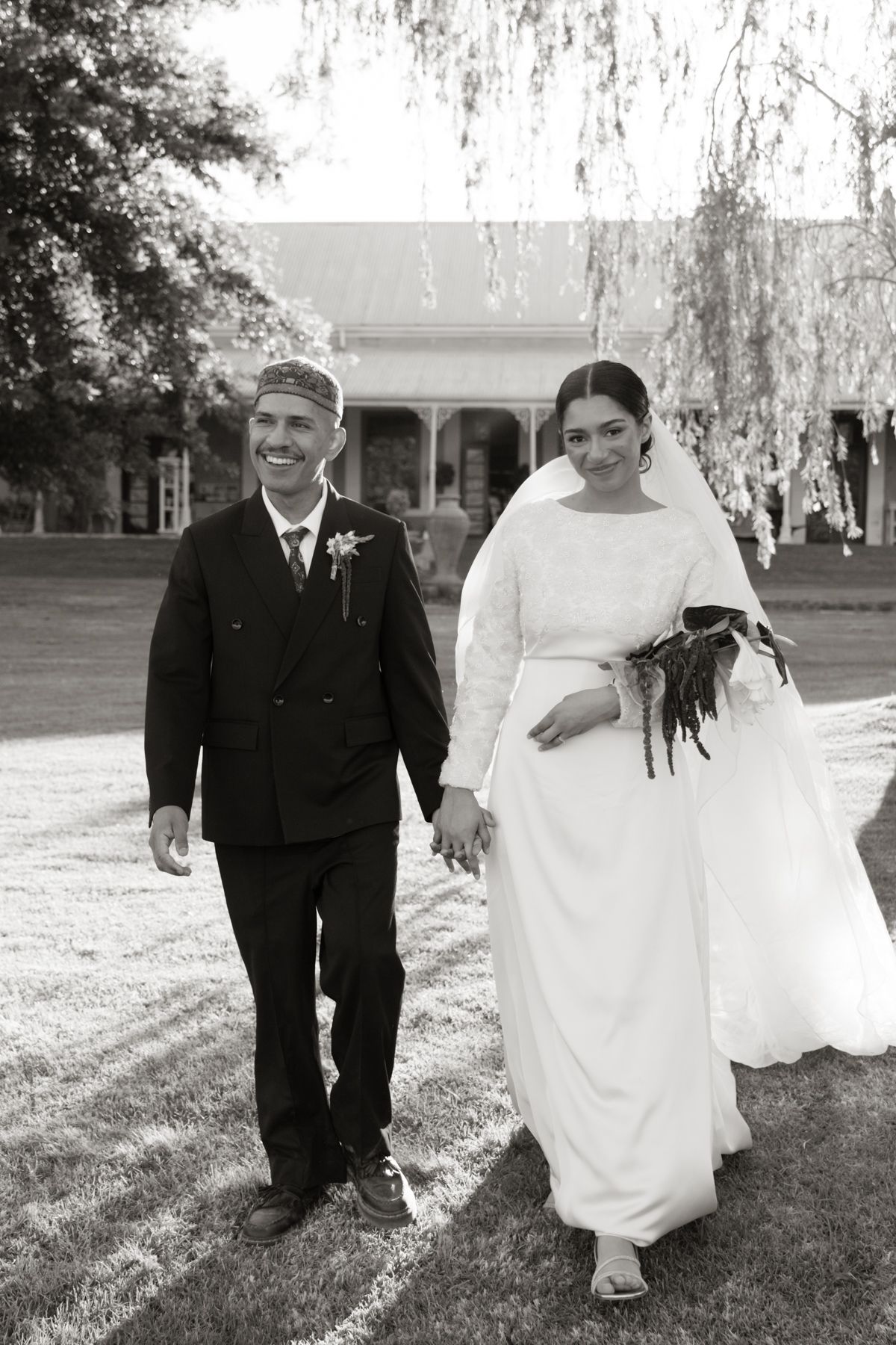 Black and white portrait of a smiling bride and groom walking hand in hand at their outdoor wedding.