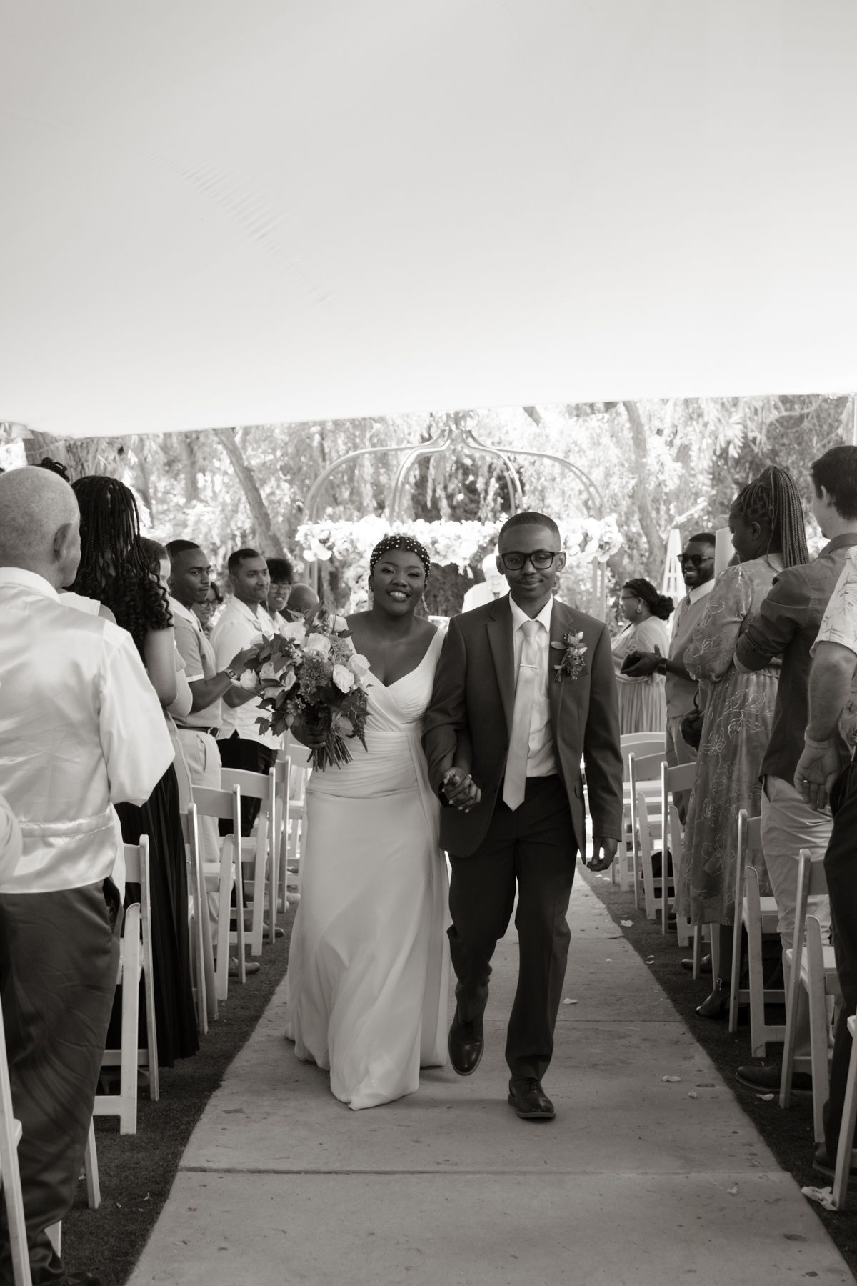 Black and white photo of newlyweds joyfully walking back down aisle after outdoor garden ceremony.