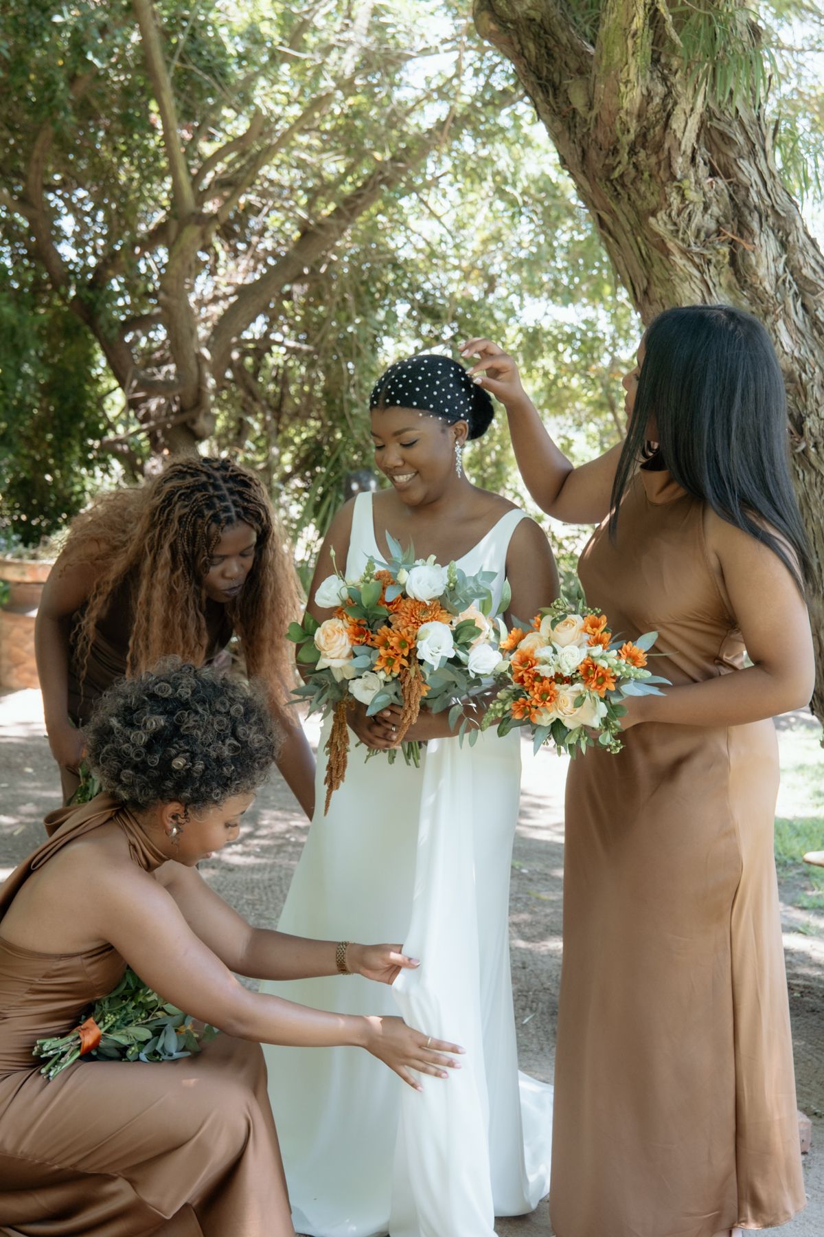 Bridesmaids in brown satin dresses help adjust bride in white gown holding orange and white bouquet outdoors.