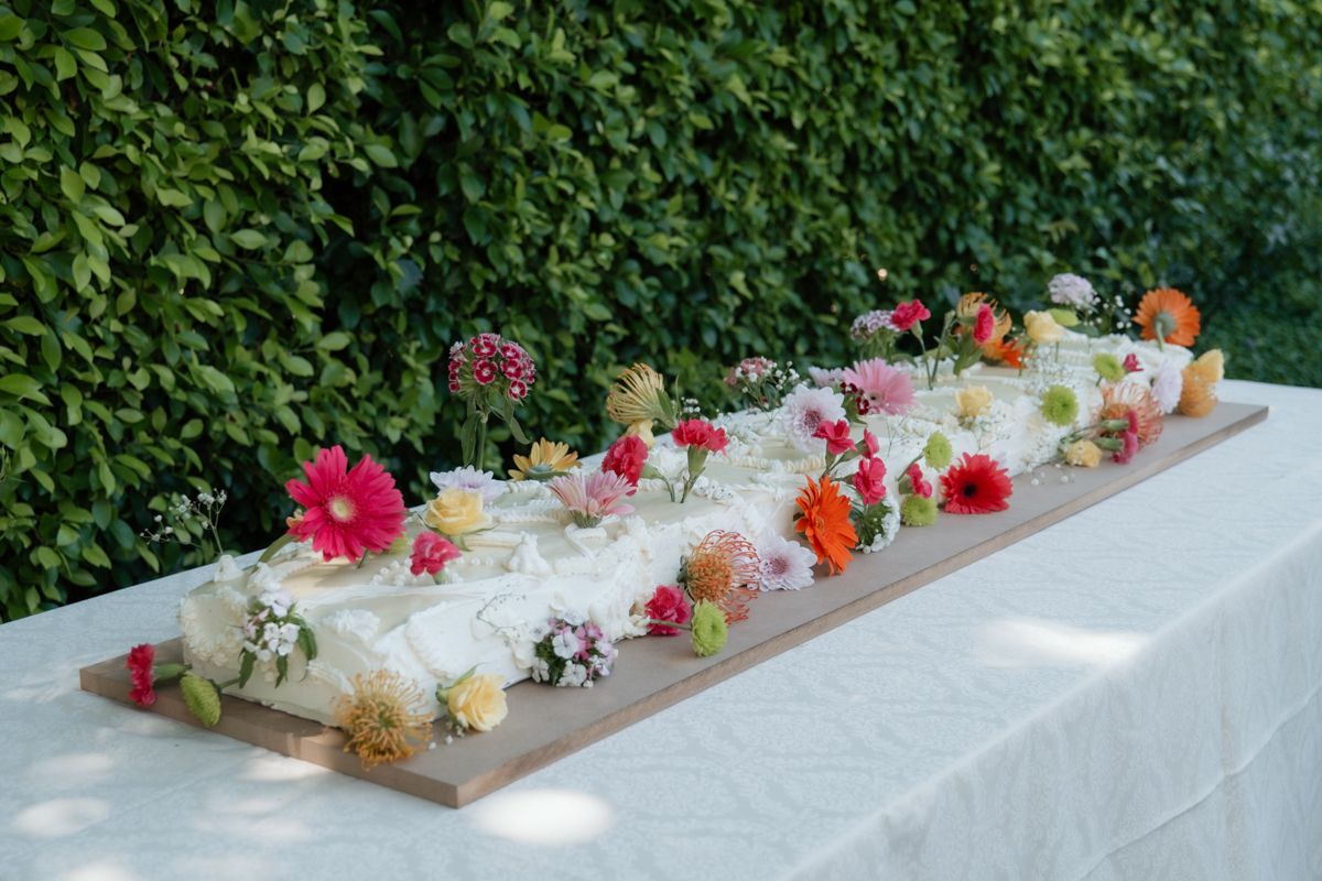 Long white sheet cake decorated with colorful fresh flowers on a white tablecloth, displayed outdoors against a green hedge.