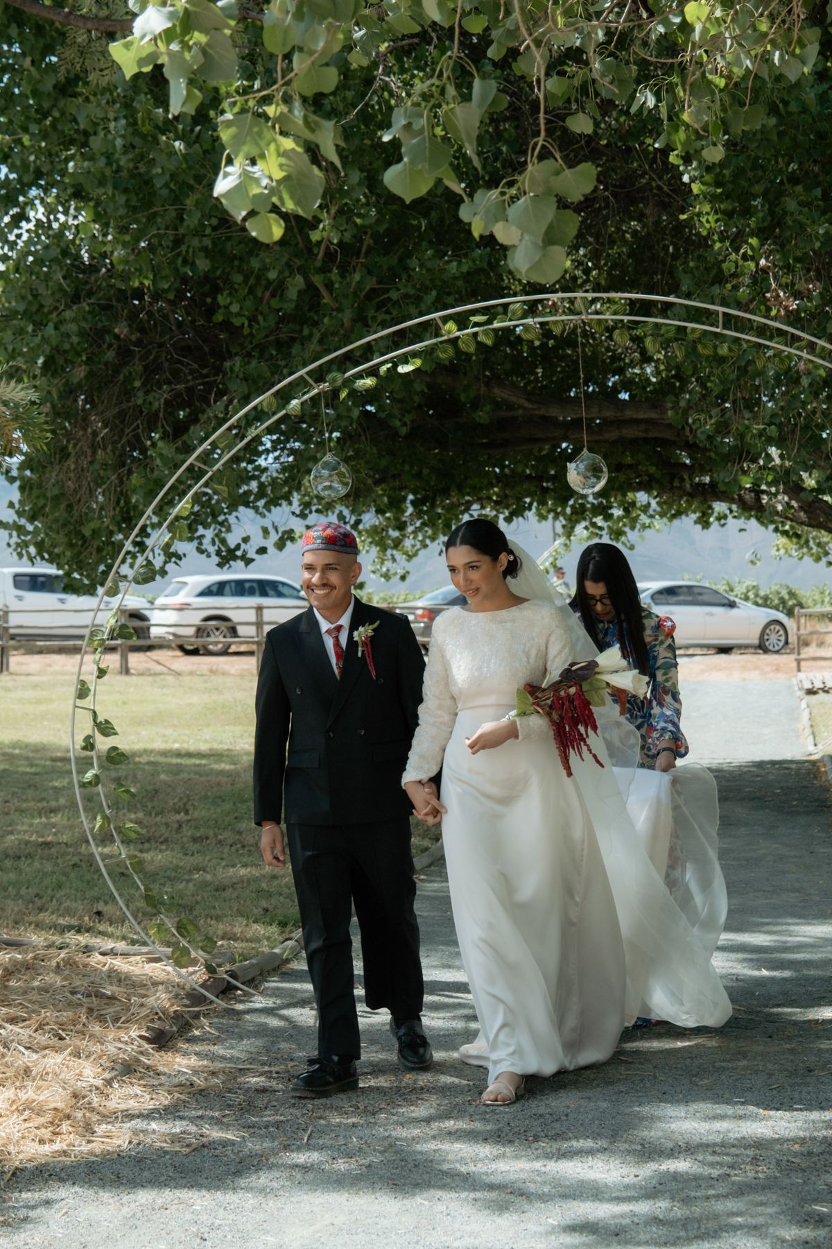 Bride in white gown walks with escort under circular arch decorated with greenery and flowers.