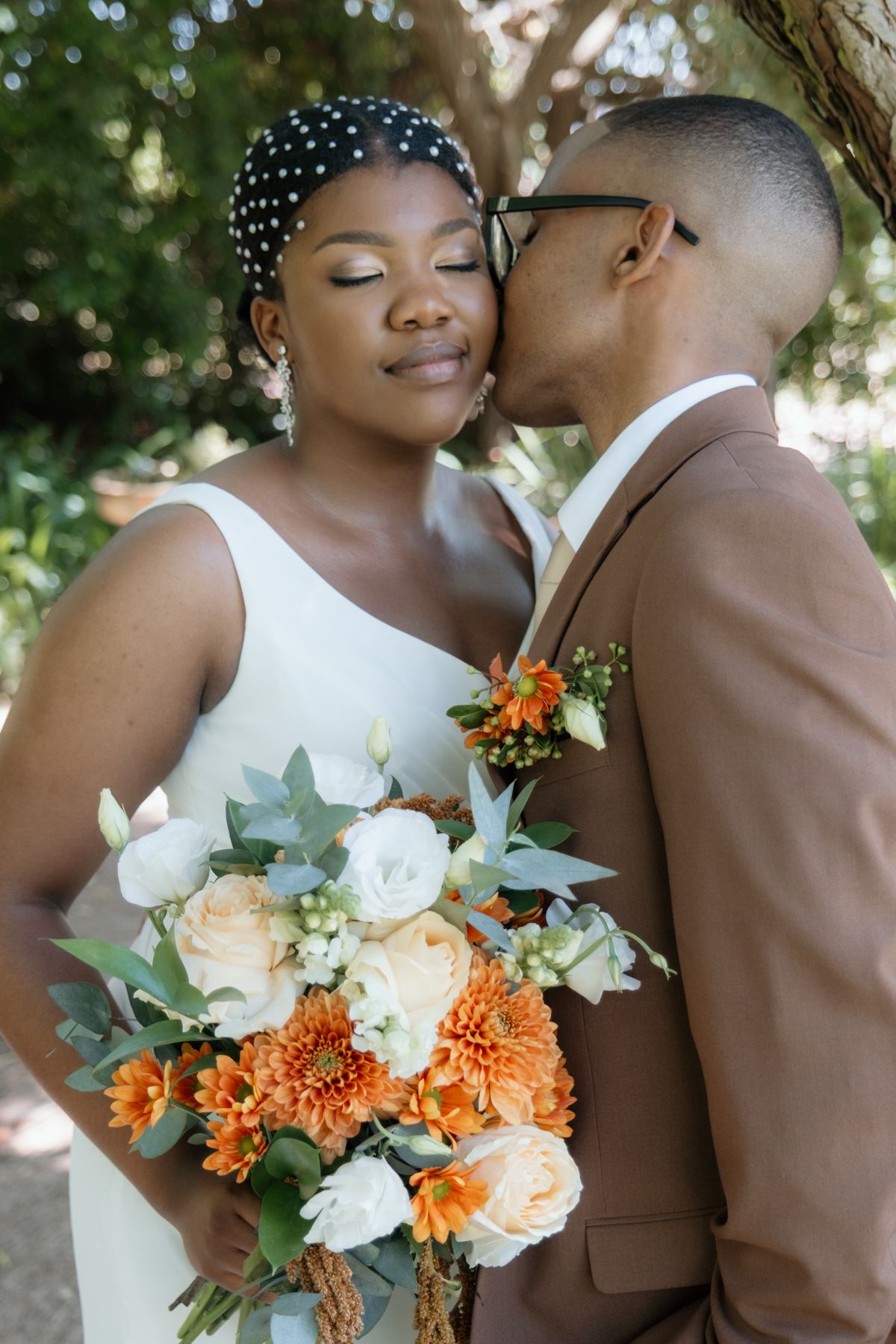 Groom in brown suit kisses bride holding orange and white bouquet under trees on their wedding day.