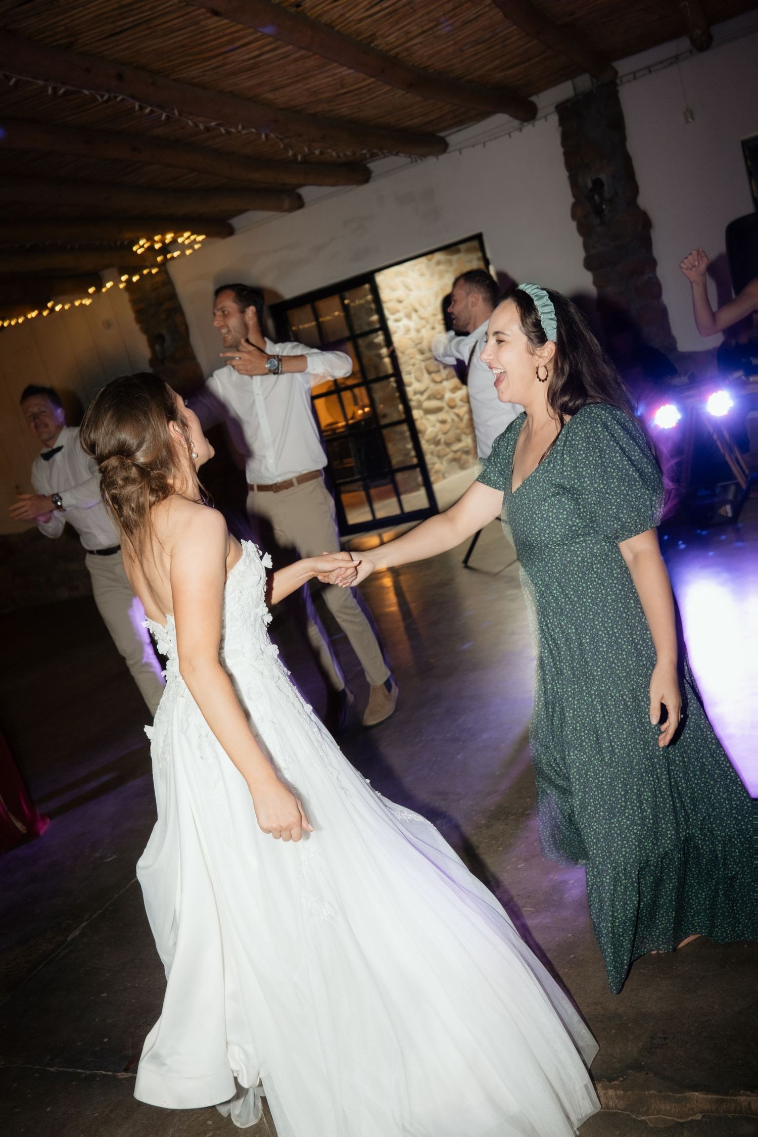 Bride in white gown dances joyfully with a guest in green dress at lively wedding reception.