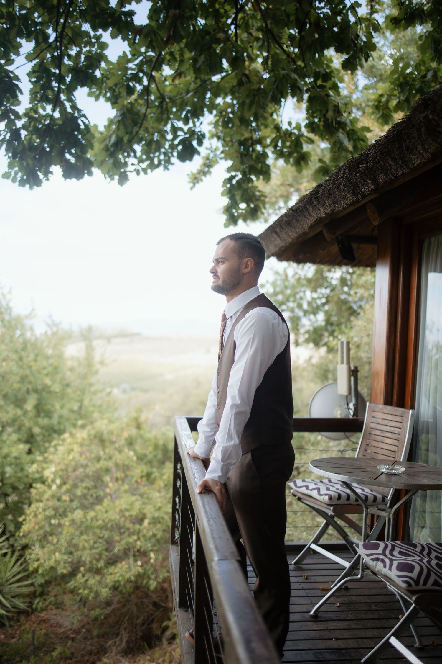 Groom in vest and tie stands on wooden balcony overlooking lush green landscape on wedding day.