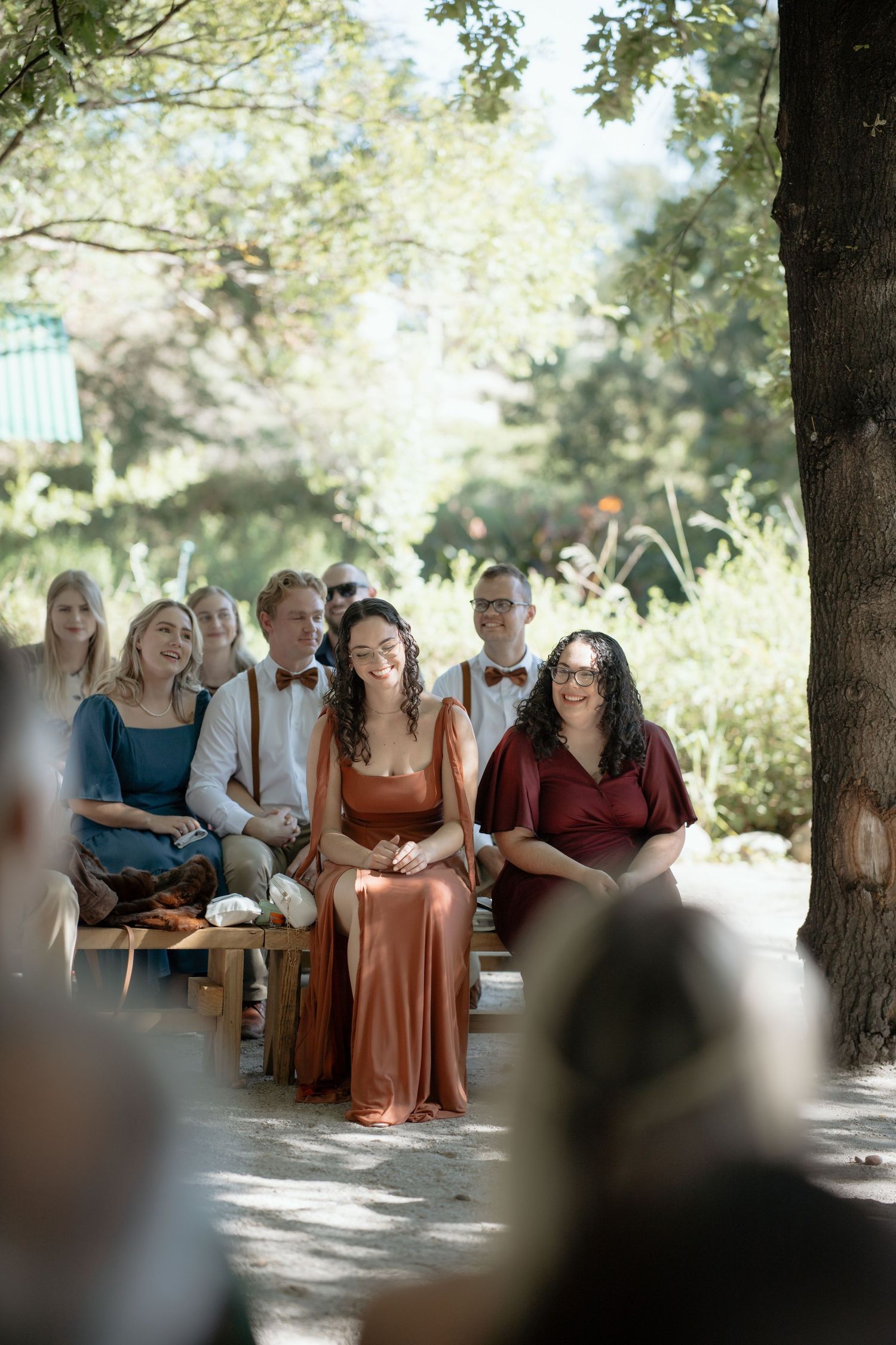 Wedding guests seated outdoors under trees smiling during ceremony, bridesmaids in rust and burgundy dresses.