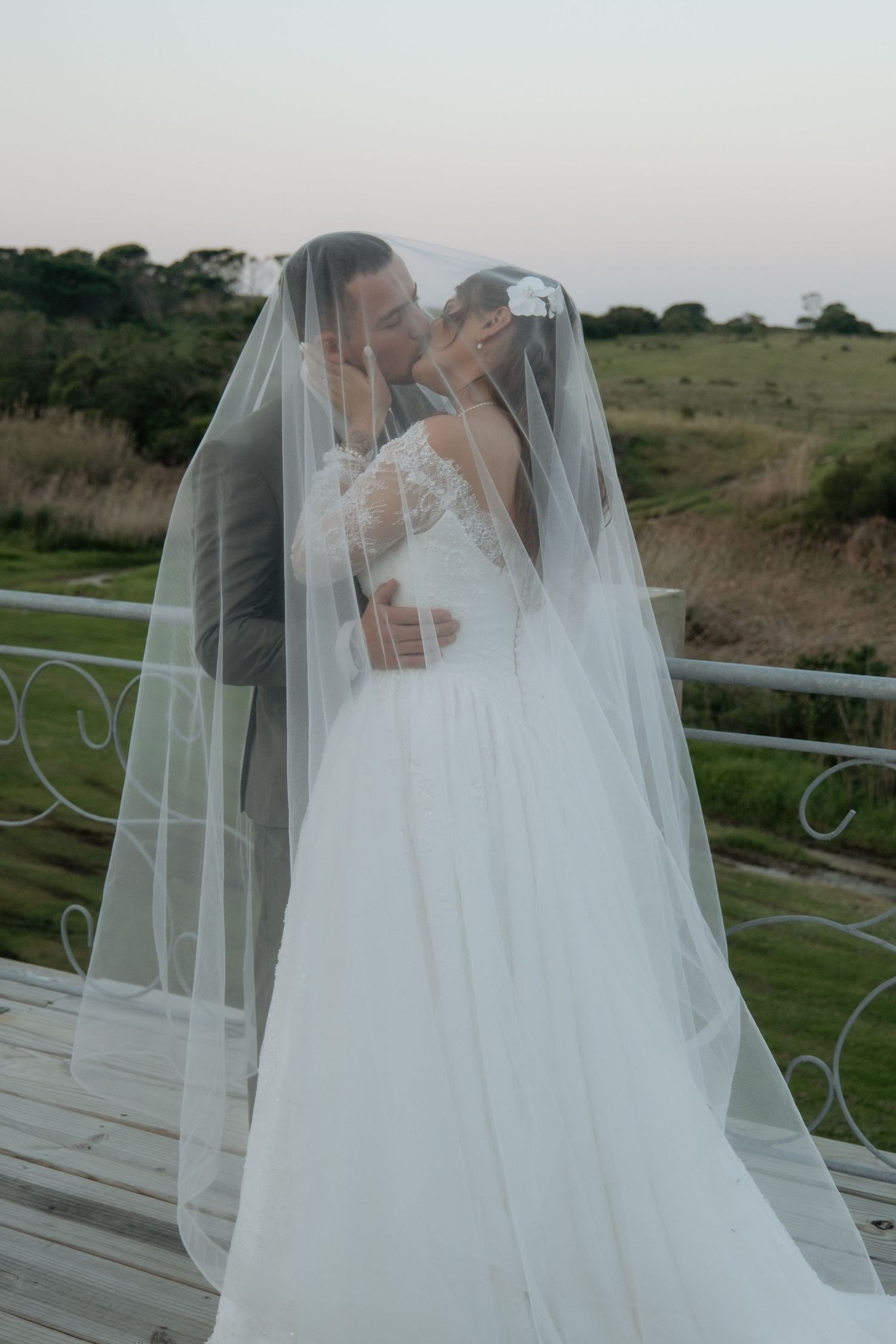 Bride and groom share romantic kiss under flowing white veil on outdoor deck with green countryside view.