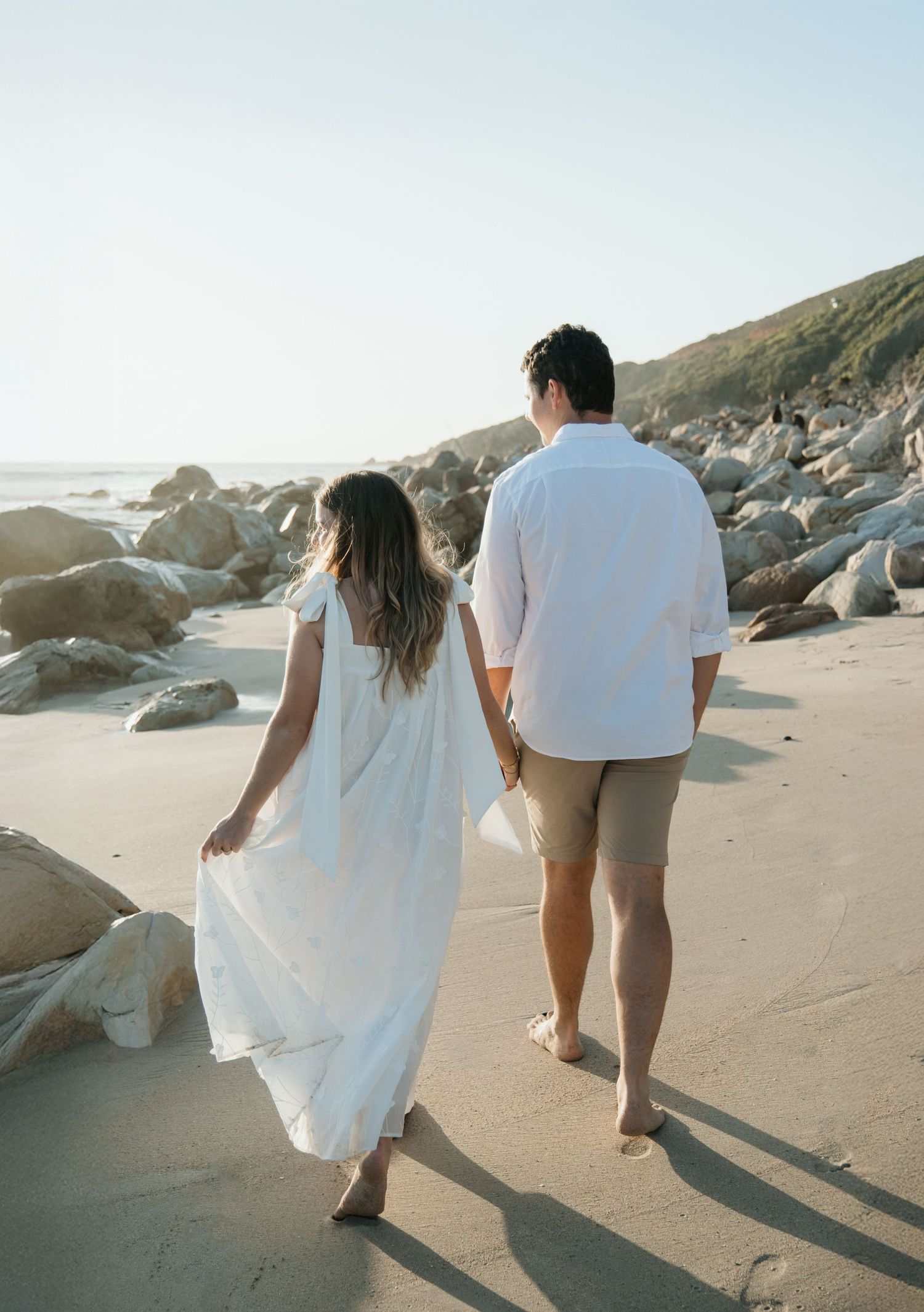 Couple walking barefoot along a rocky beach at sunset in white attire on the Kogel Bay Shoreline