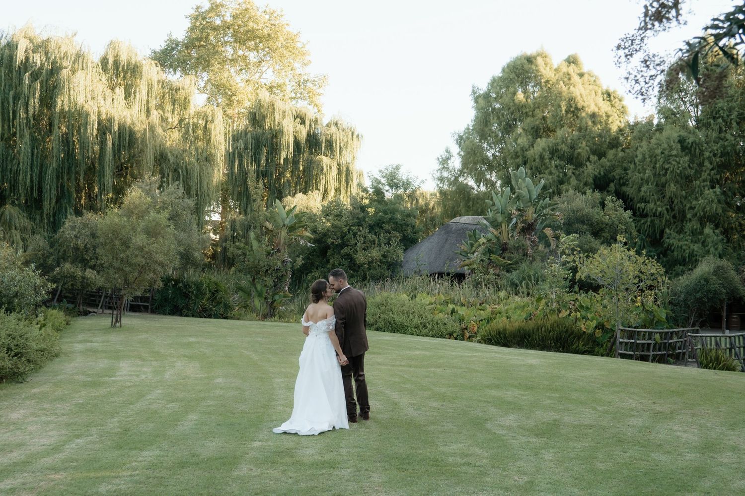 Bride and groom stand together on lush green lawn surrounded by willow trees and gardens at outdoor wedding venue.