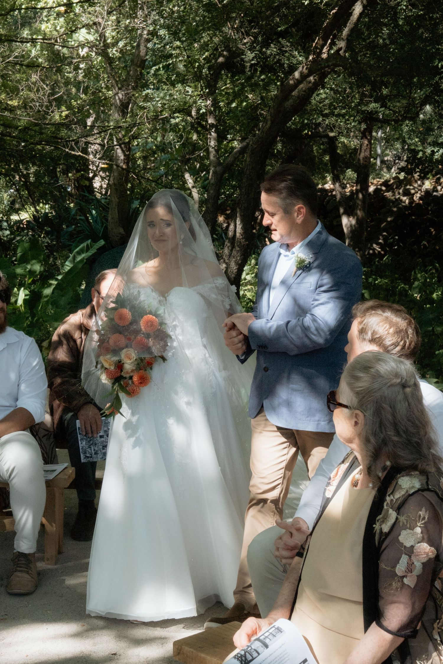 Bride in white gown and veil holds colorful bouquet beside groom in blue suit at outdoor wedding ceremony.