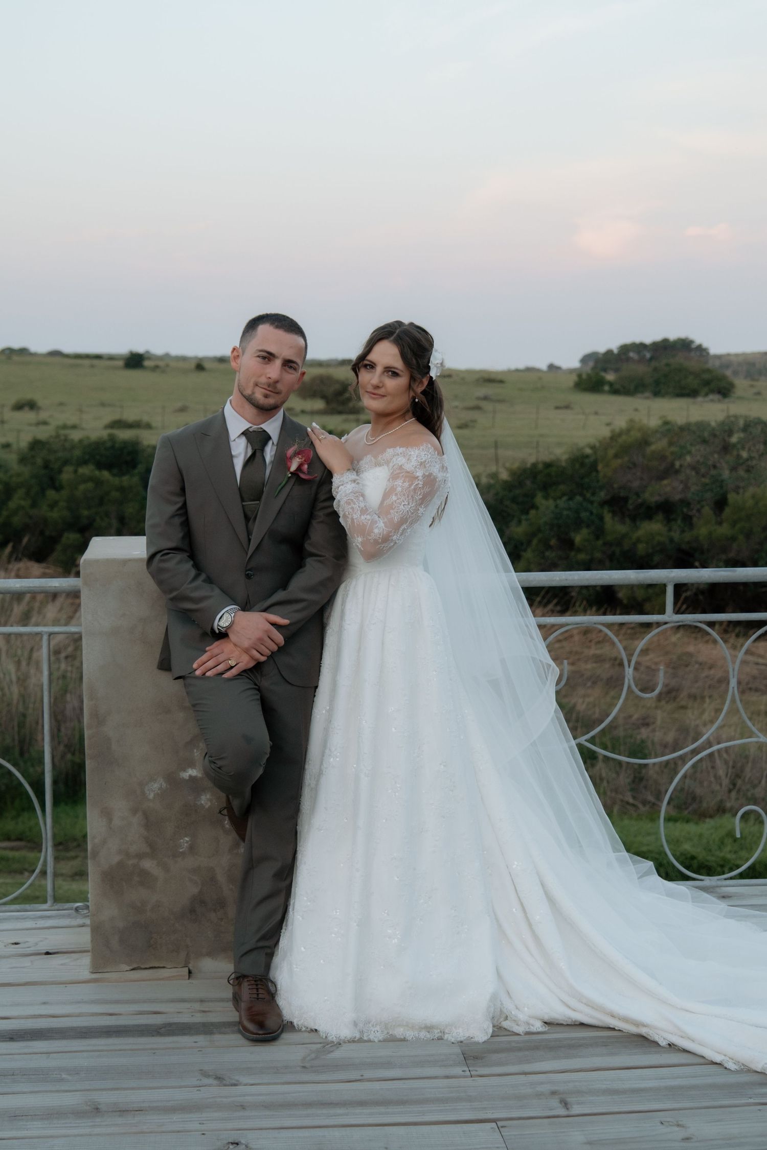 Bride and groom posing on balcony at sunset with rolling green fields behind them.
