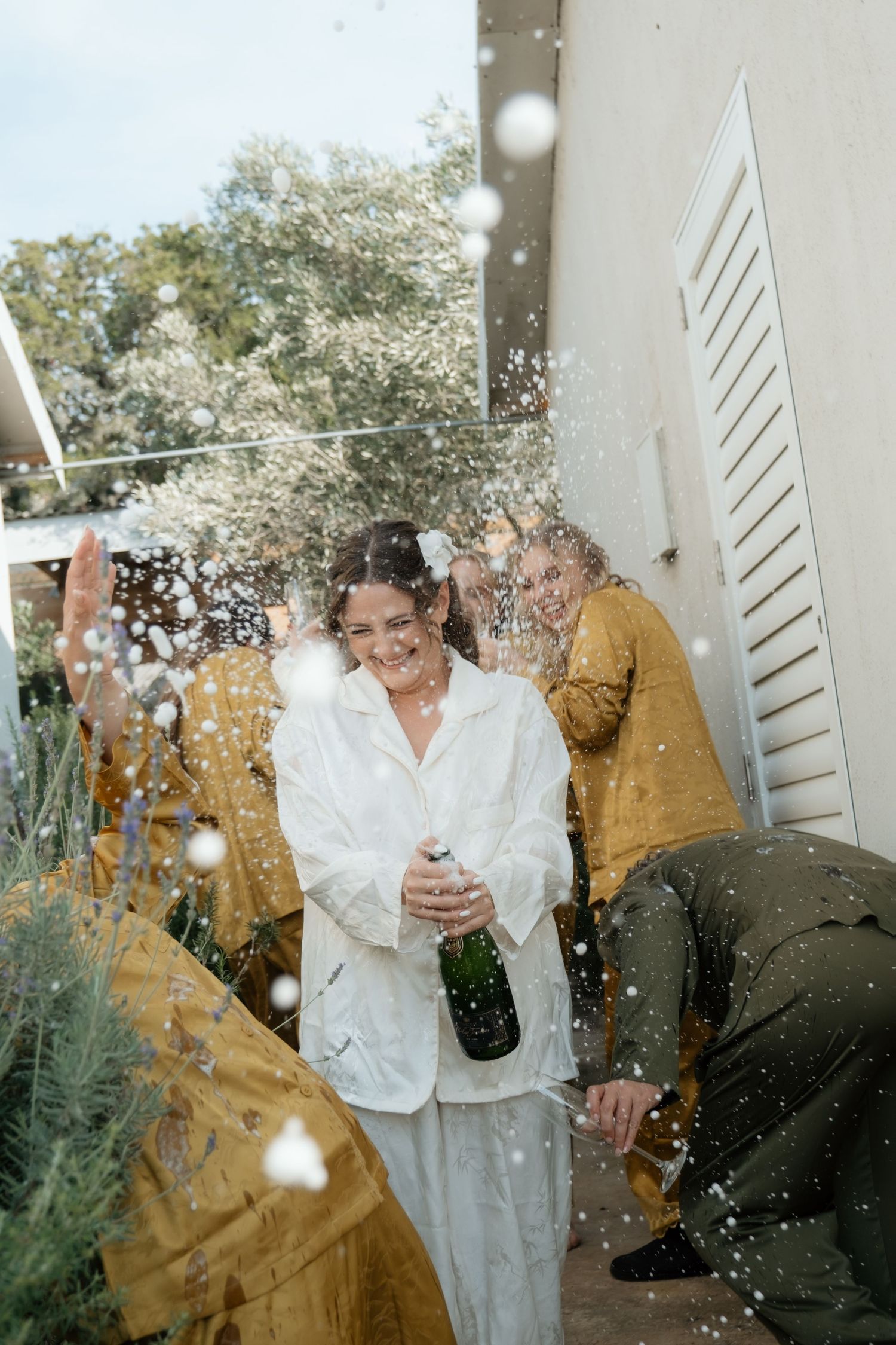 Woman in white outfit joyfully popping champagne bottle with friends celebrating outdoors.