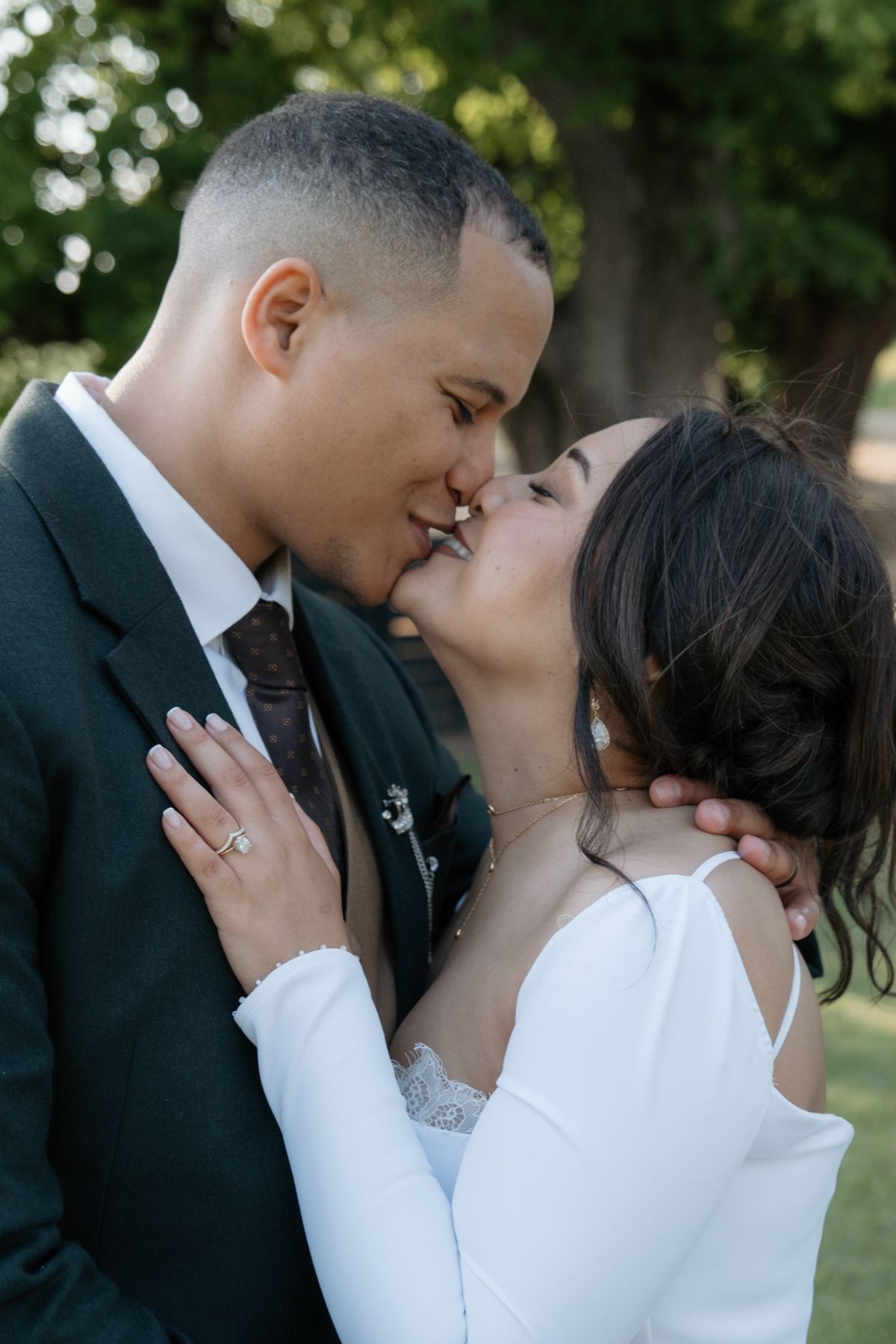 A bride and groom share a romantic kiss outdoors surrounded by lush green trees.