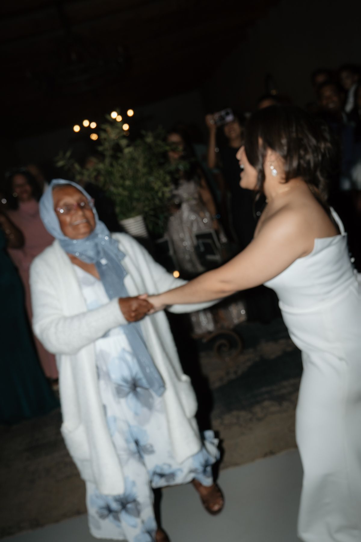 Bride in white gown joyfully dancing and holding hands with elderly woman in blue floral wrap at reception.