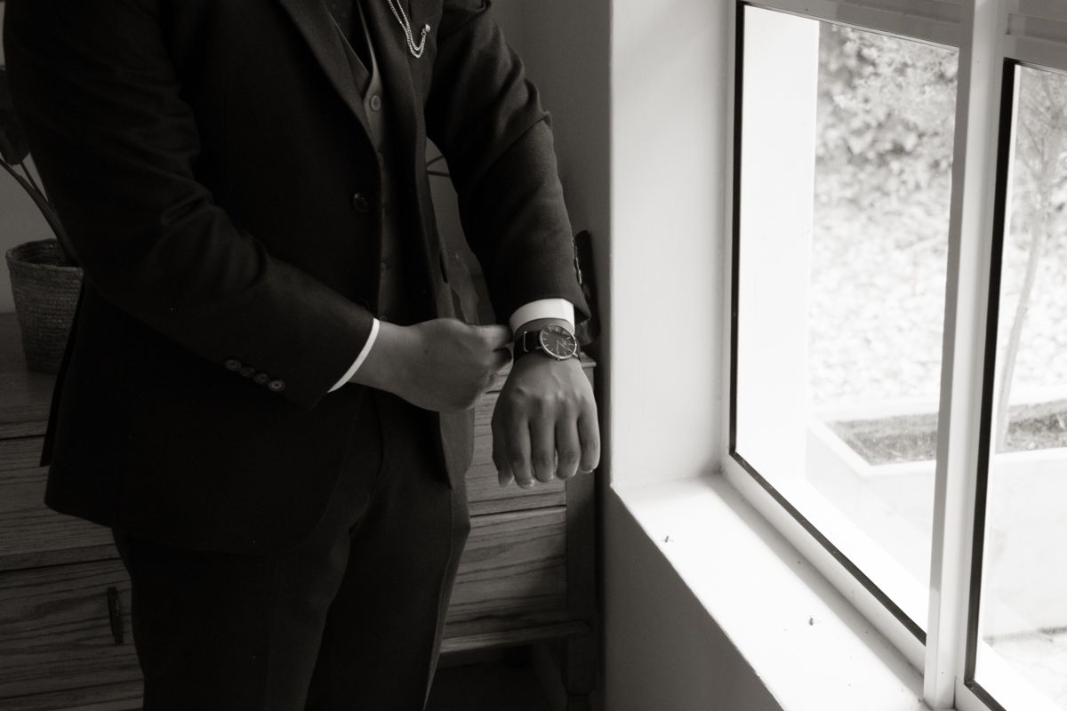Close-up black and white photo of groom adjusting his watch cufflinks by a bright window.
