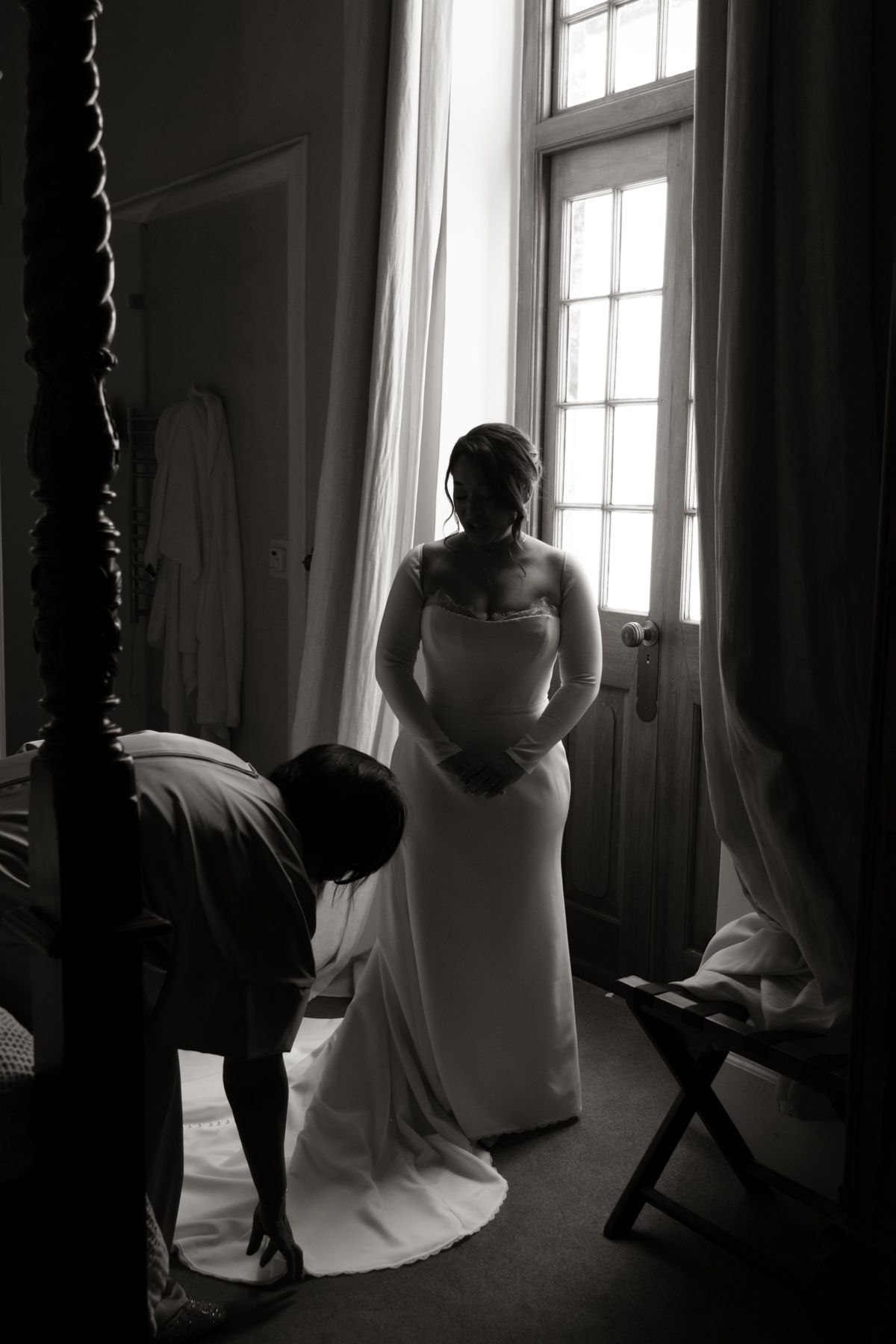 Black and white silhouette of bride in wedding gown being dressed near bright window before ceremony.
