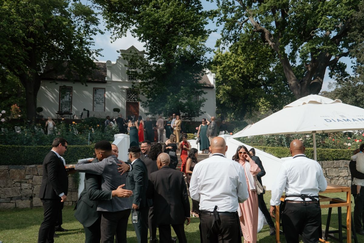 Wedding guests mingle on garden lawn outside elegant estate venue on sunny day.