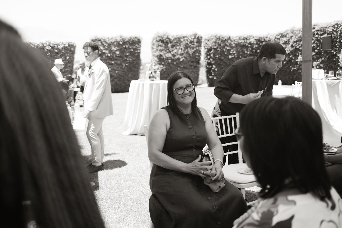 Black and white photo of a smiling woman in glasses sitting outdoors at an elegant garden event.