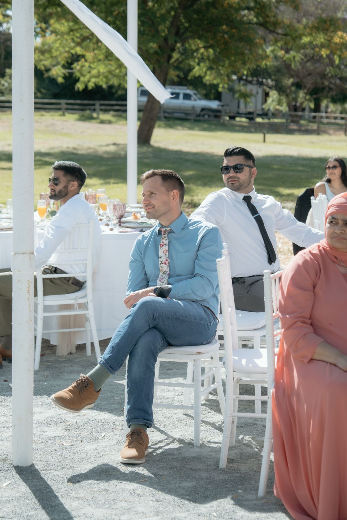 Wedding guests seated outdoors at sunny reception, man in blue shirt holds wine glass.