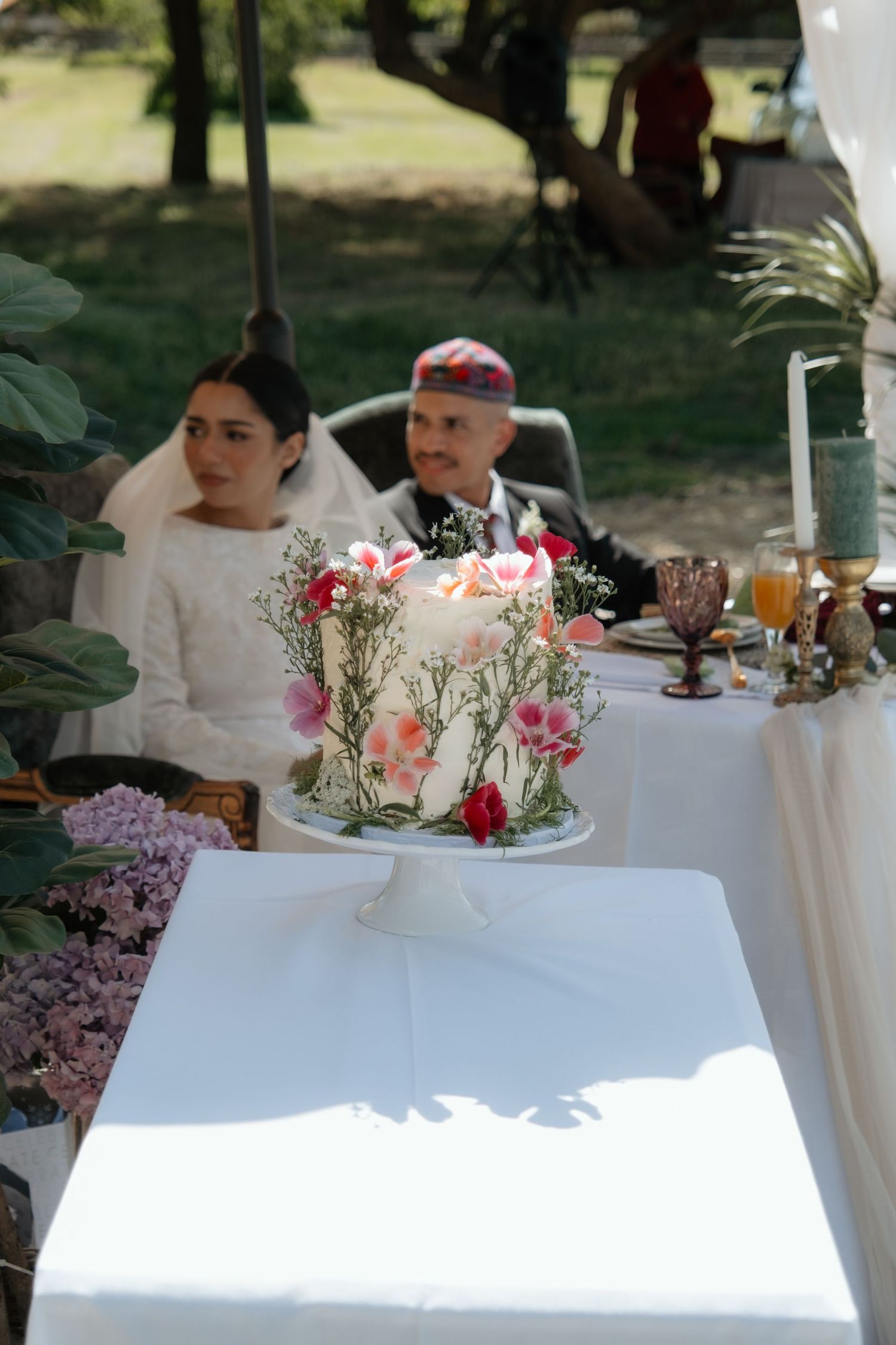 Elegant floral wedding cake with pink blooms displayed at outdoor reception table with bride and groom.