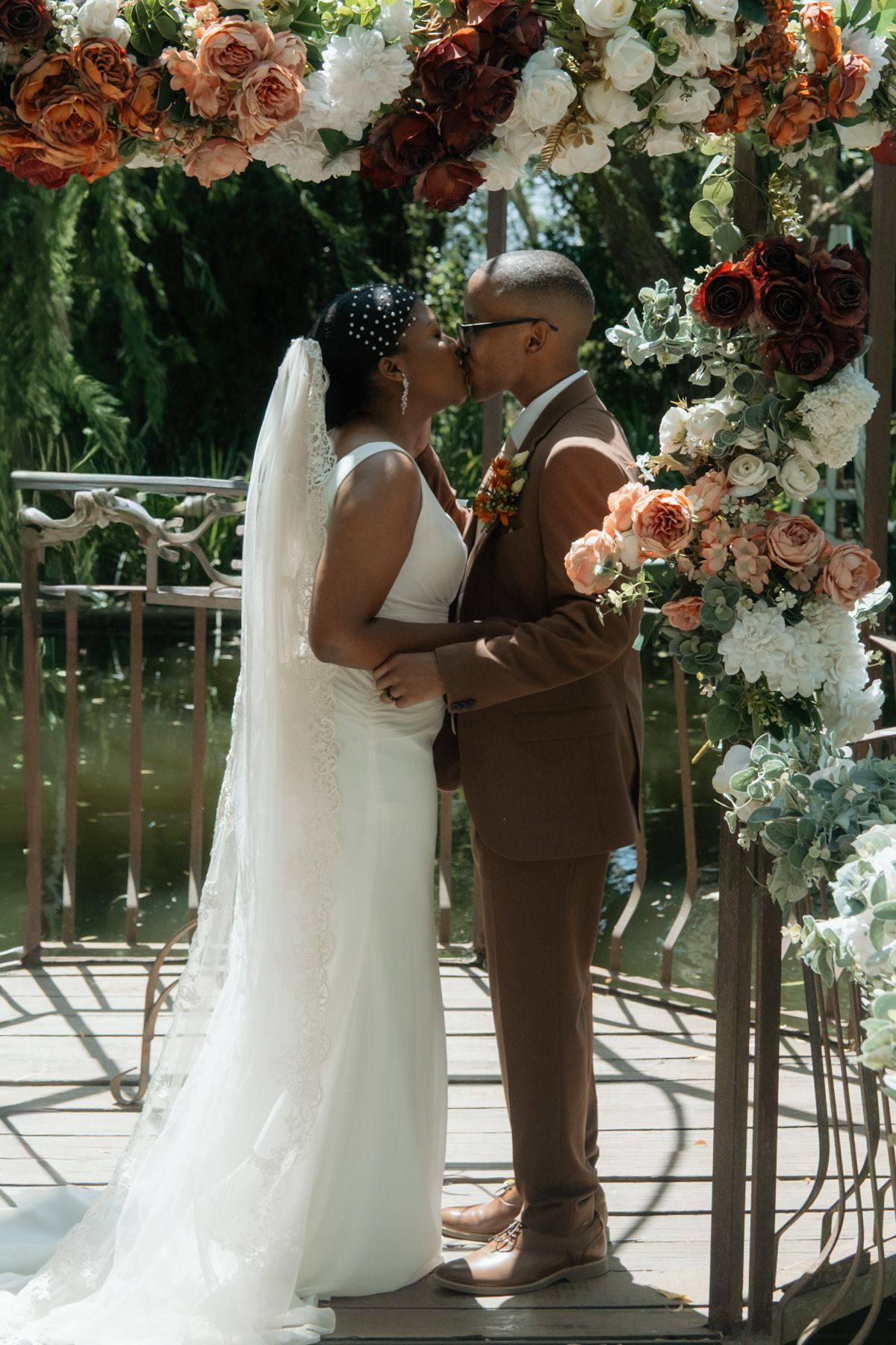 Bride and groom share first kiss under floral arch with roses at outdoor waterside wedding.