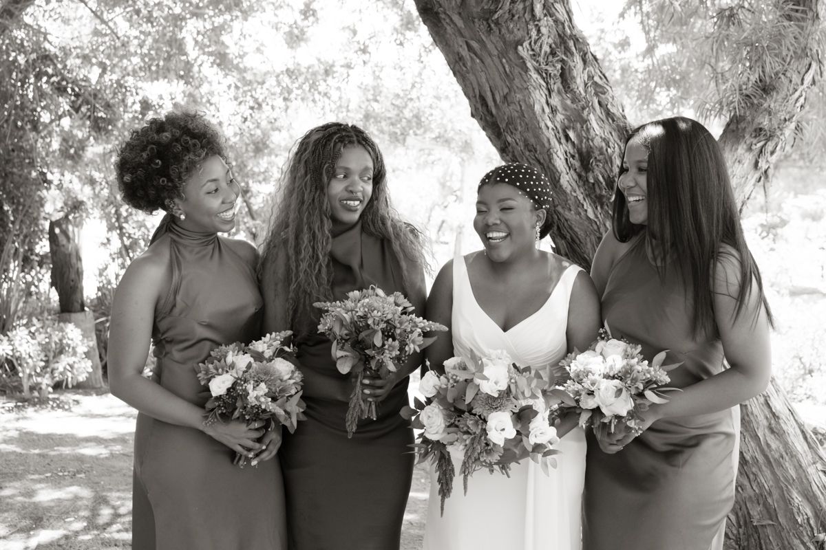 Black and white photo of bride in white gown laughing with three bridesmaids holding floral bouquets under a tree.
