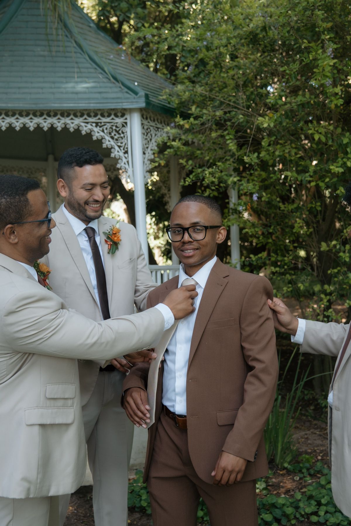 Two men adjust young boys brown suit tie while groomsman smiles near garden gazebo.